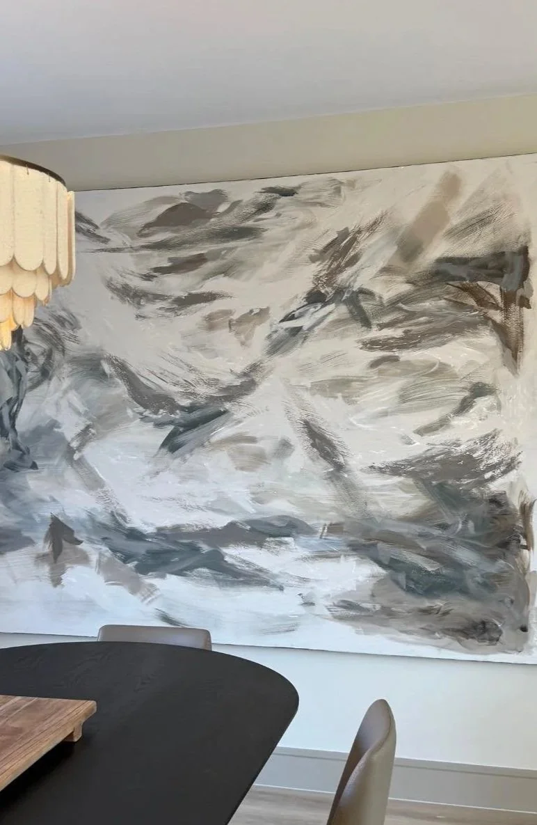 Abstract black, white, and gray painting on a wall in a dining room with a dark table and light-colored chairs.