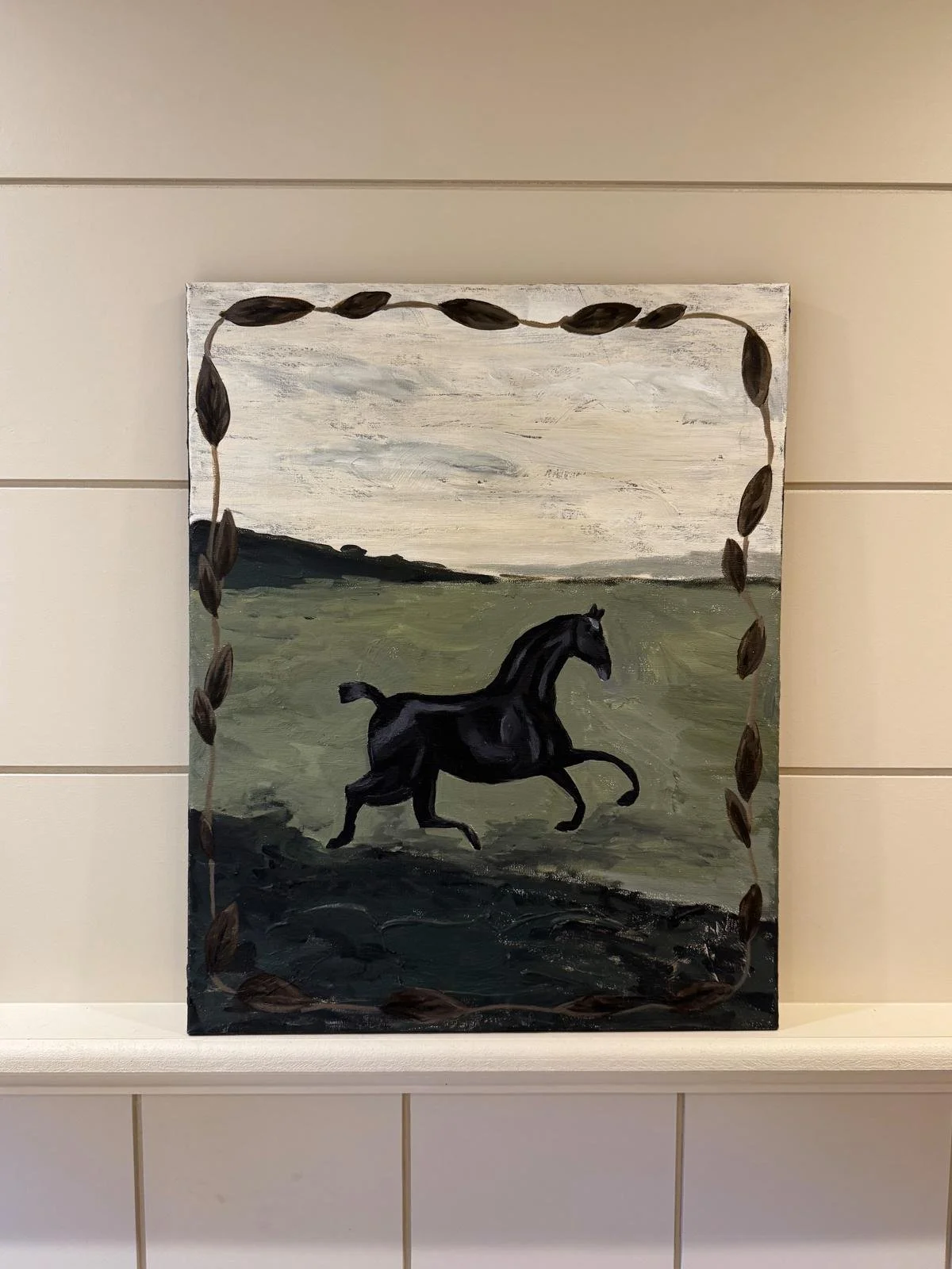 Painting of a black horse running through a green field with a cloudy sky in the background, framed with painted dark leaves.