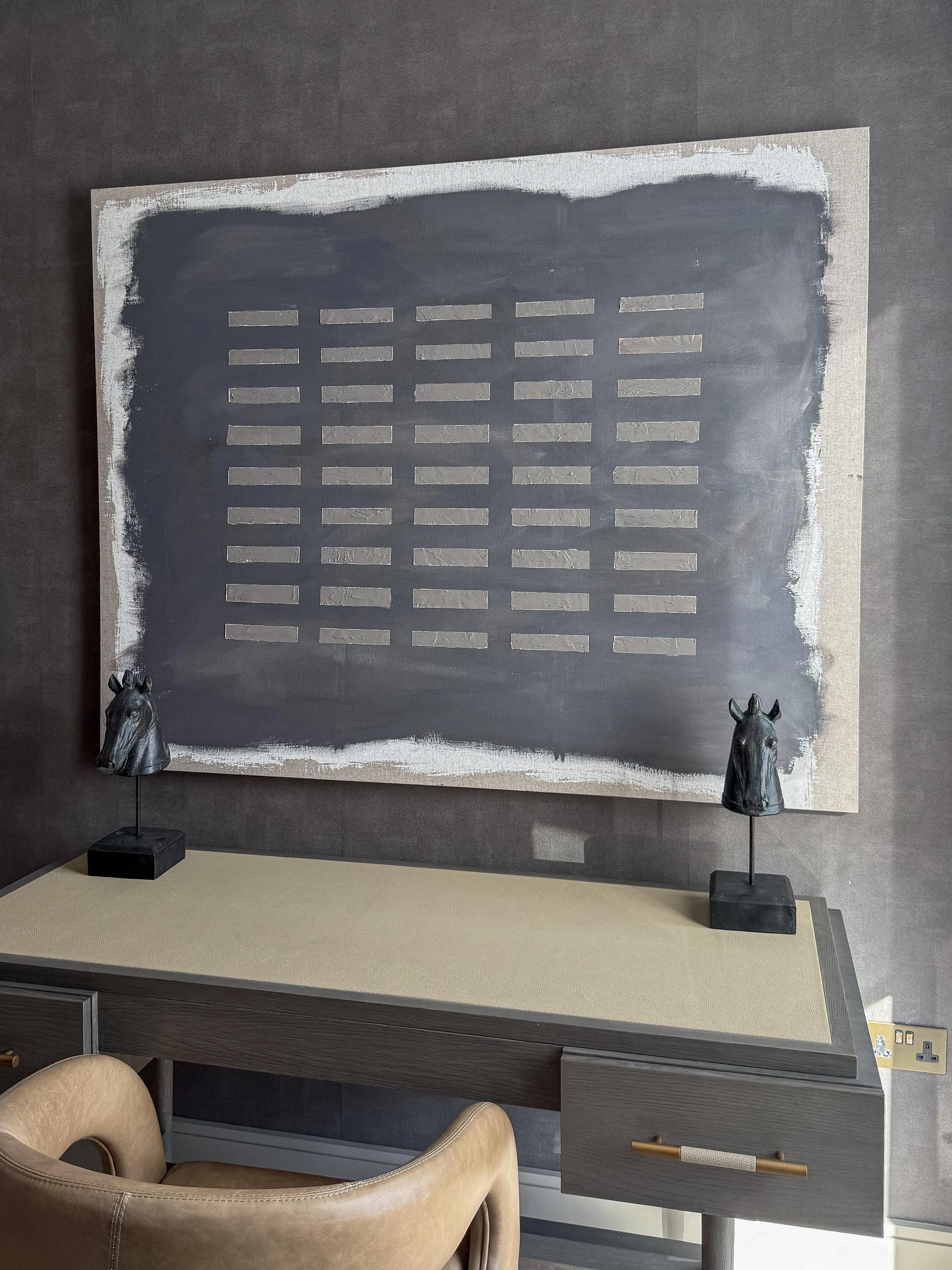 Decorative wall art featuring a grid of metallic rectangles on a dark background, flanked by two small horse head sculptures on stands, placed on a beige surface with a dark wood cabinet below.