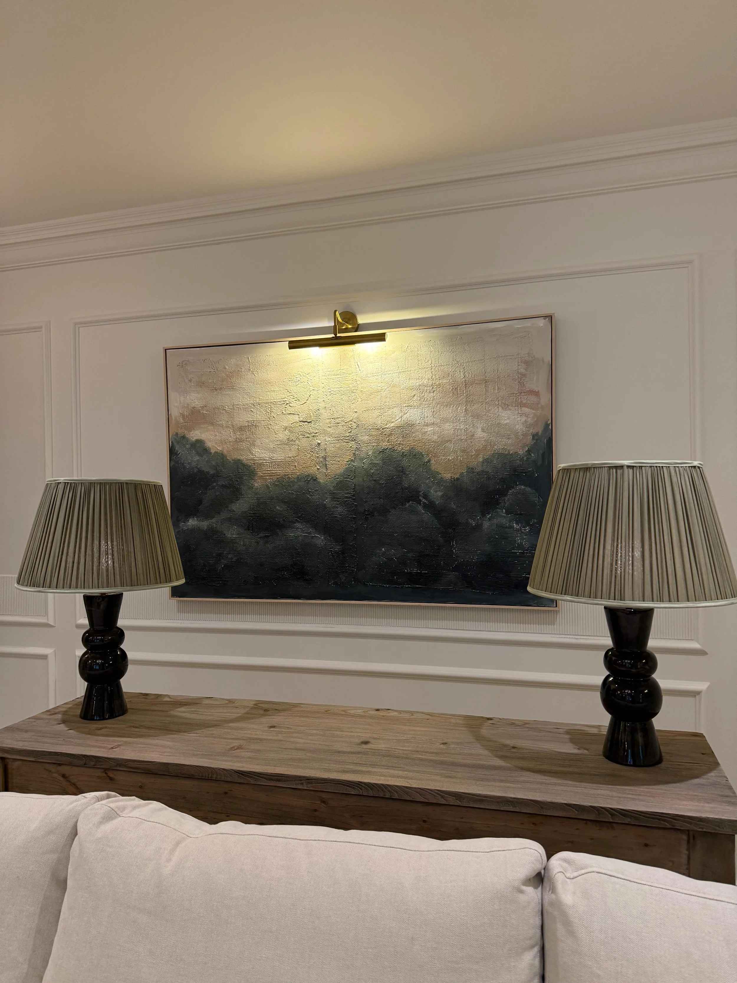 A decorated interior wall with a painting illuminated by a picture light, flanked by two table lamps on a wooden console table, and a white sofa in the foreground.