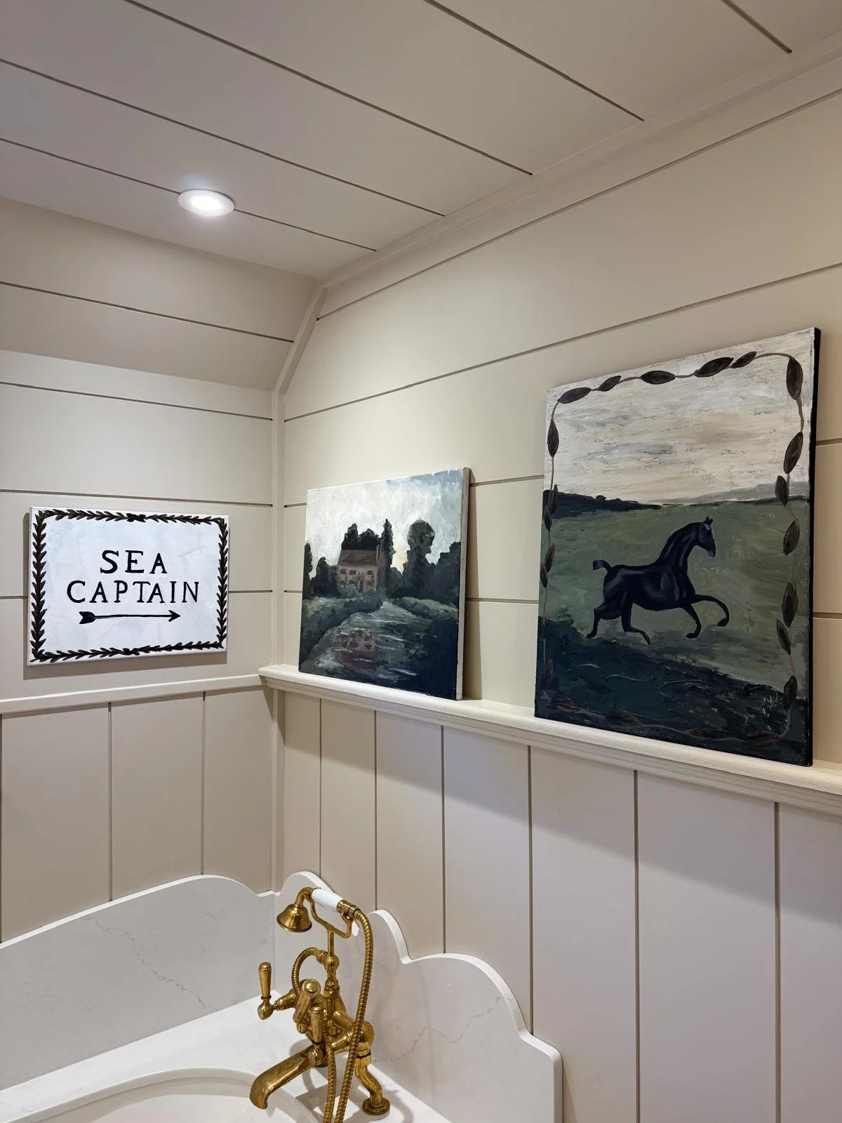 A corner of a bathroom with beige paneled walls, a gold faucet, and three paintings on a shelf. One painting has a barn, the second depicts a landscape, and the third shows a black horse running in fields. A sign on the wall reads "Sea Captain" with an arrow.