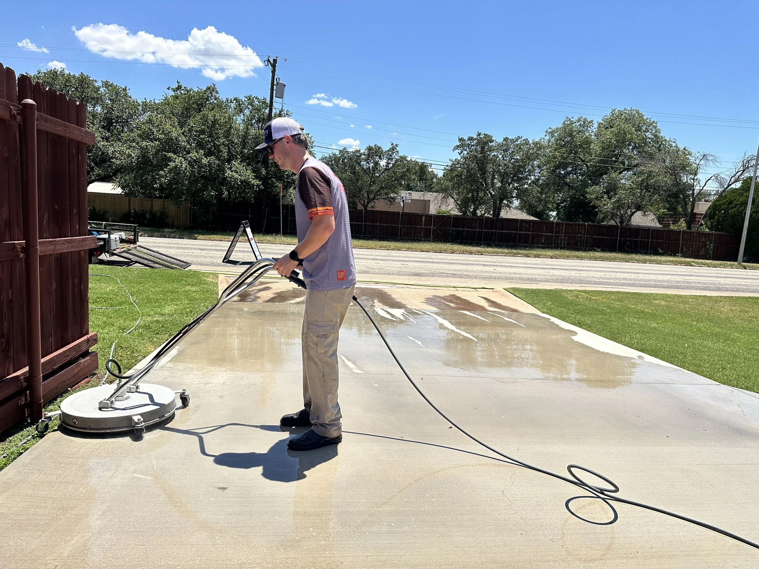 A man is pressure washing the concrete porch outside a house on a cloudy day. He is wearing a gray shirt, tan pants, and a cap. There are trees, parked cars, and a cloudy sky in the background.
