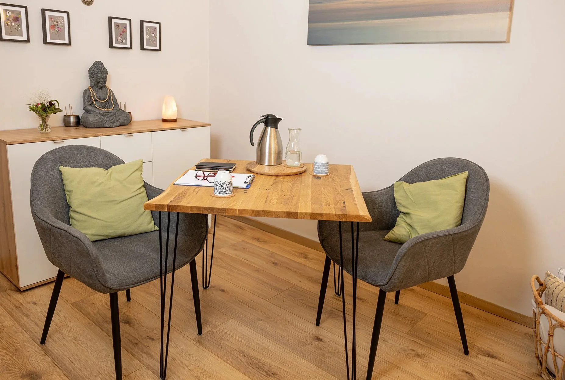 Miriam Wiese Praxistisch, an dem Gespräche stattfinden. Teekanne mit Wasserflasche und Becher, Notizblock und Brille liegen auf dem Tisch, 2 Stühle stehen daran. Dahinter eine Kommode mit einem sitzenden buddah mit Gebetskette und Räucherstäbchen.