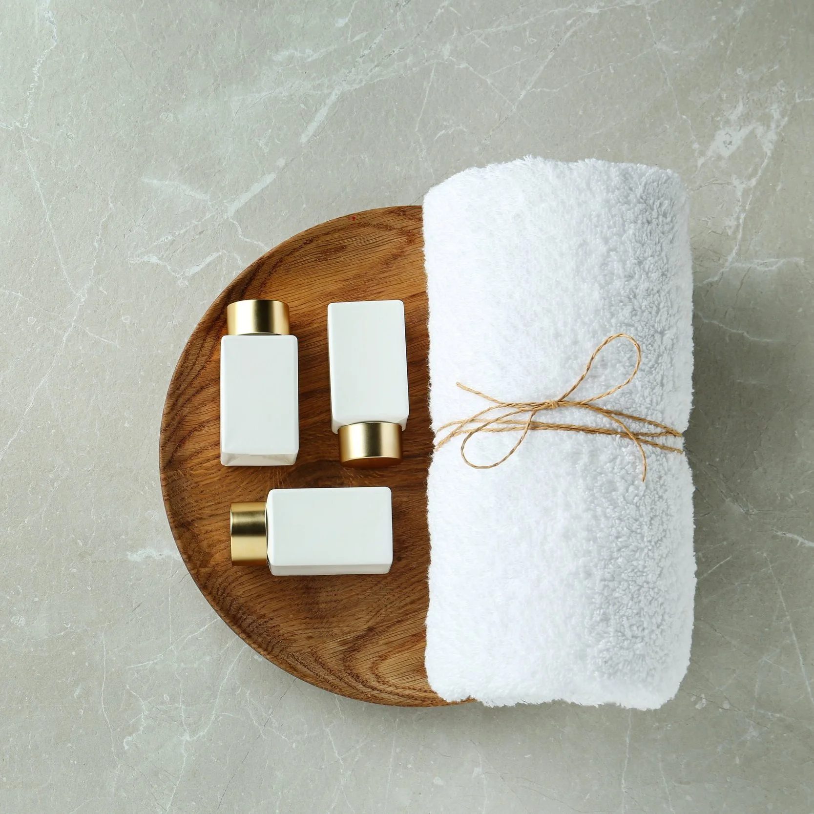 White rolled towel tied with a brown string, placed on a wooden plate with three white bottles with gold caps beside it, on a light gray marble surface.