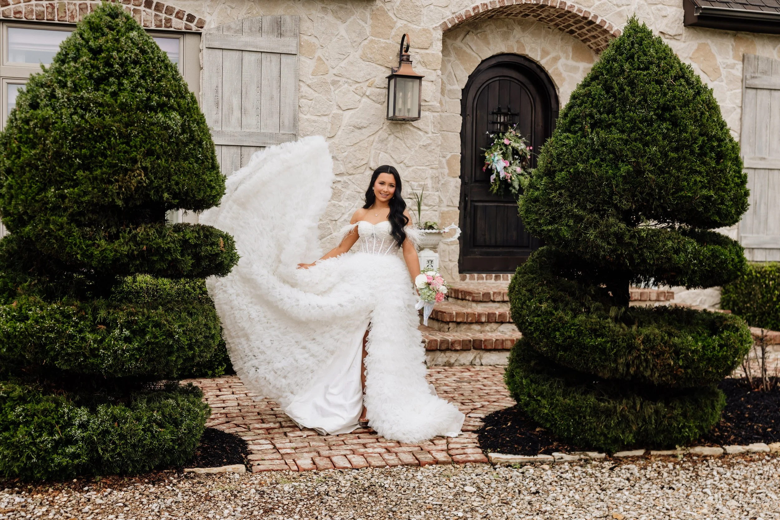 A bride in a white wedding dress with a long train and feathered sleeves, holding a flower bouquet, standing in front of a house with a stone and brick exterior, surrounded by greenery and bushes.