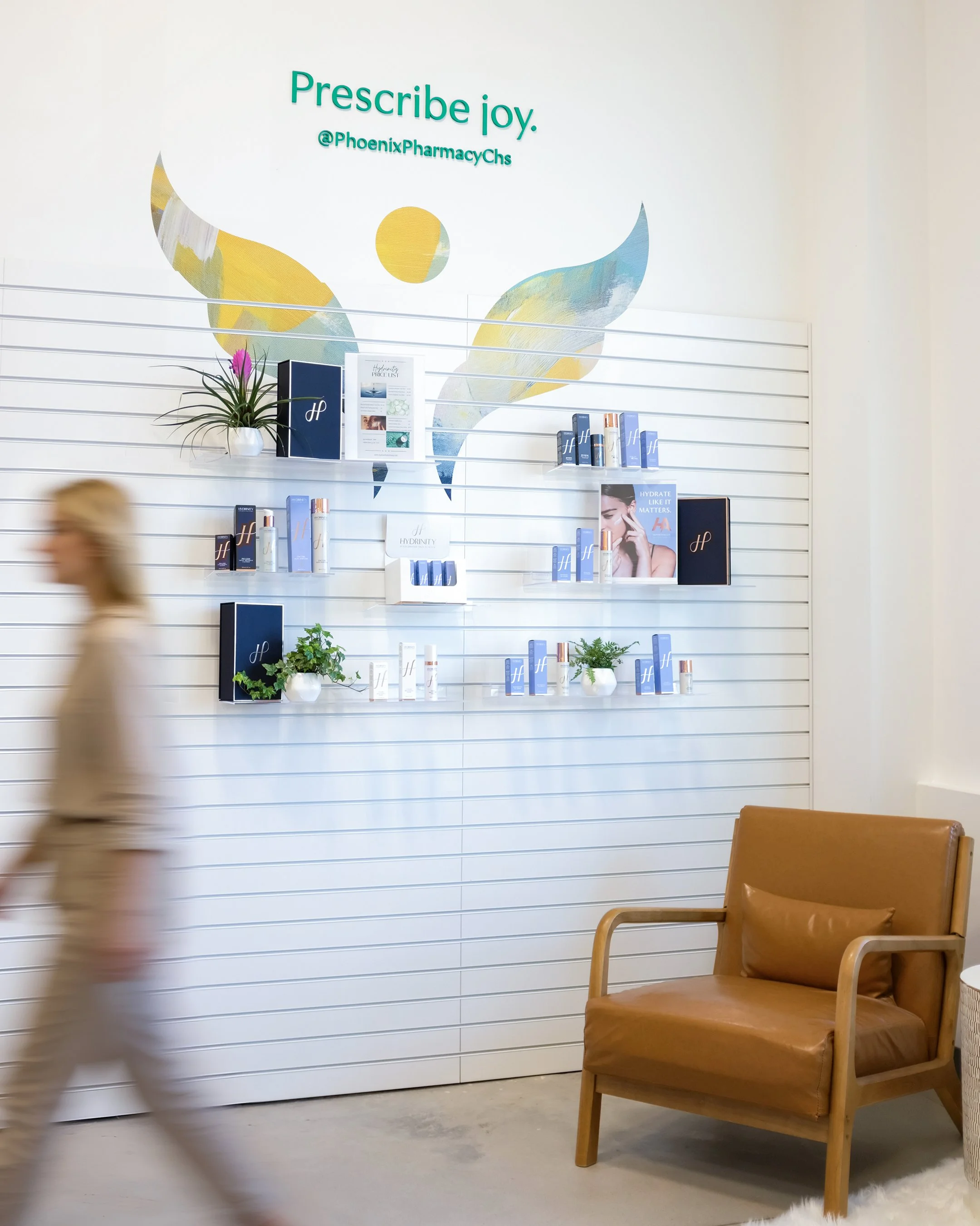 A retail display wall with skincare or beauty products, plants, and promotional signage, with a wooden armchair nearby and a person walking past.