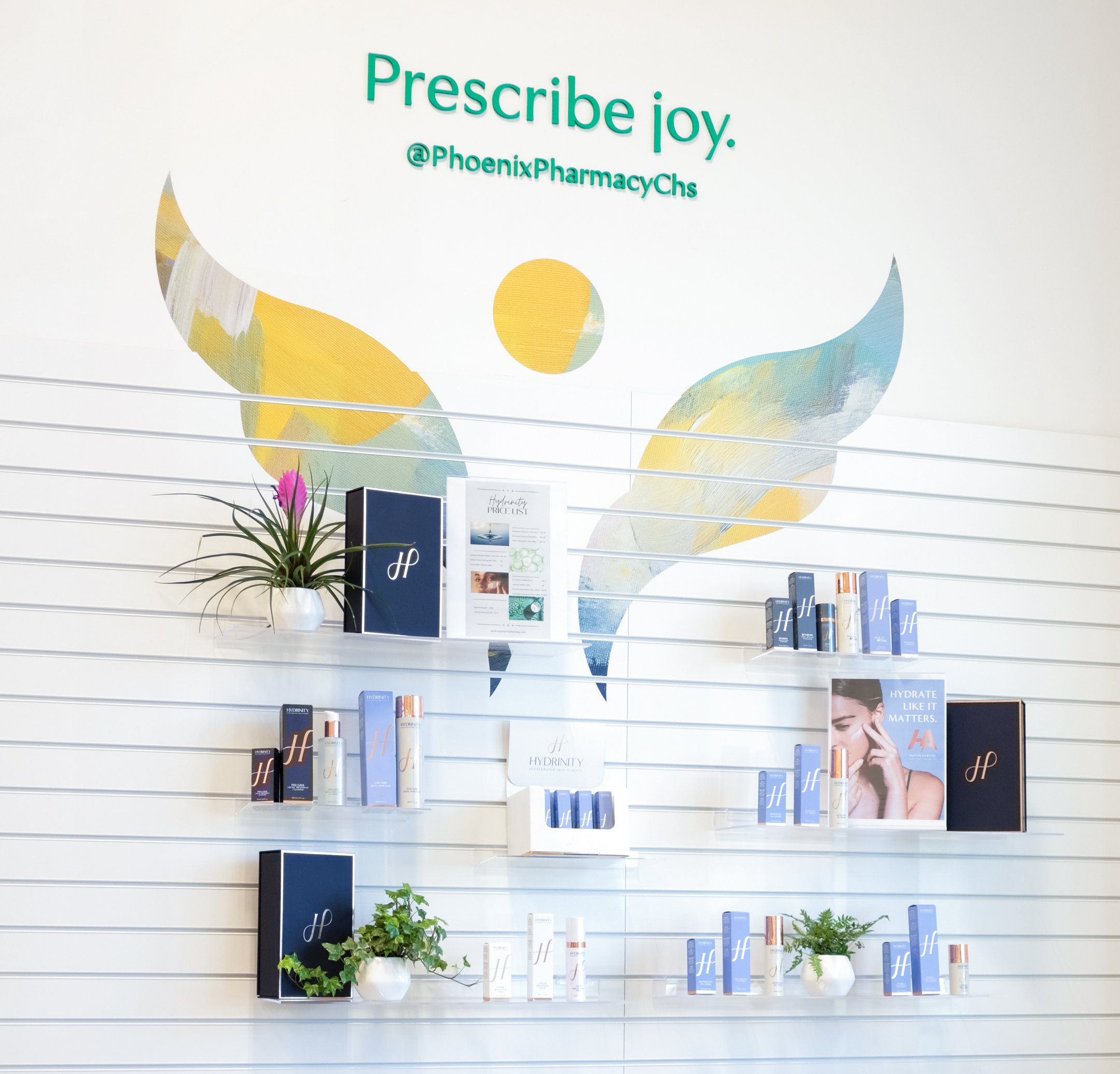 Display of skincare or beauty products with a plant and promotional signage on a wall with a colorful angel wing and circle artwork, and the text 'Prescribe joy. @PhoenixPharmacyChs'.