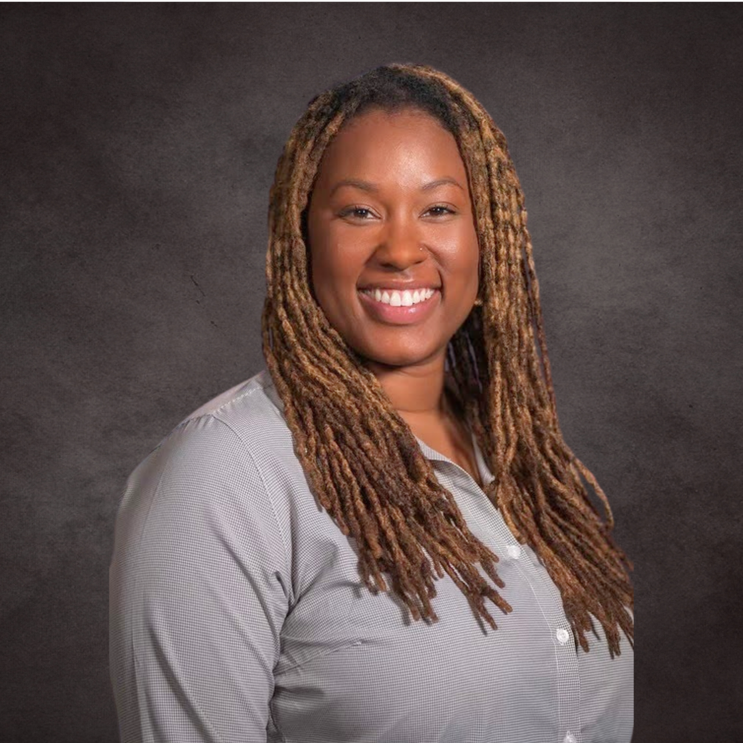 Portrait of a smiling woman with field dreadlocks wearing a light gray button-up shirt against a dark background.