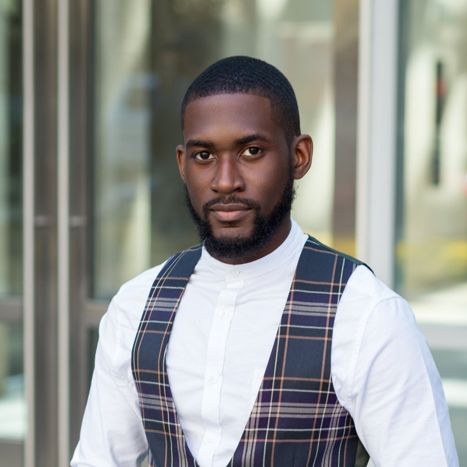 Portrait of a young man with a beard and short hair, wearing a white button-up shirt and a plaid vest, standing outside in front of a building with glass windows.