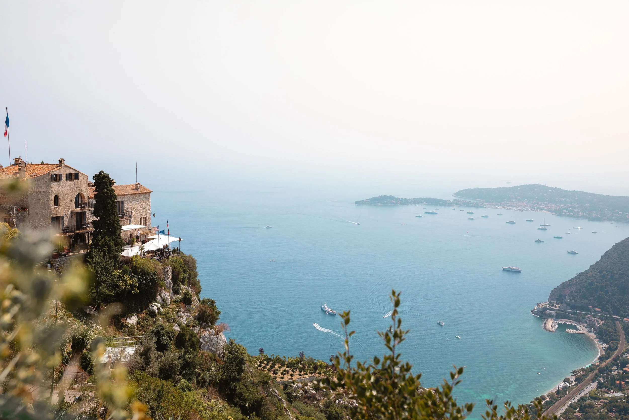 A scenic view of a coastal area with houses on a hillside, boats on the water, and a distant shoreline under a hazy sky.