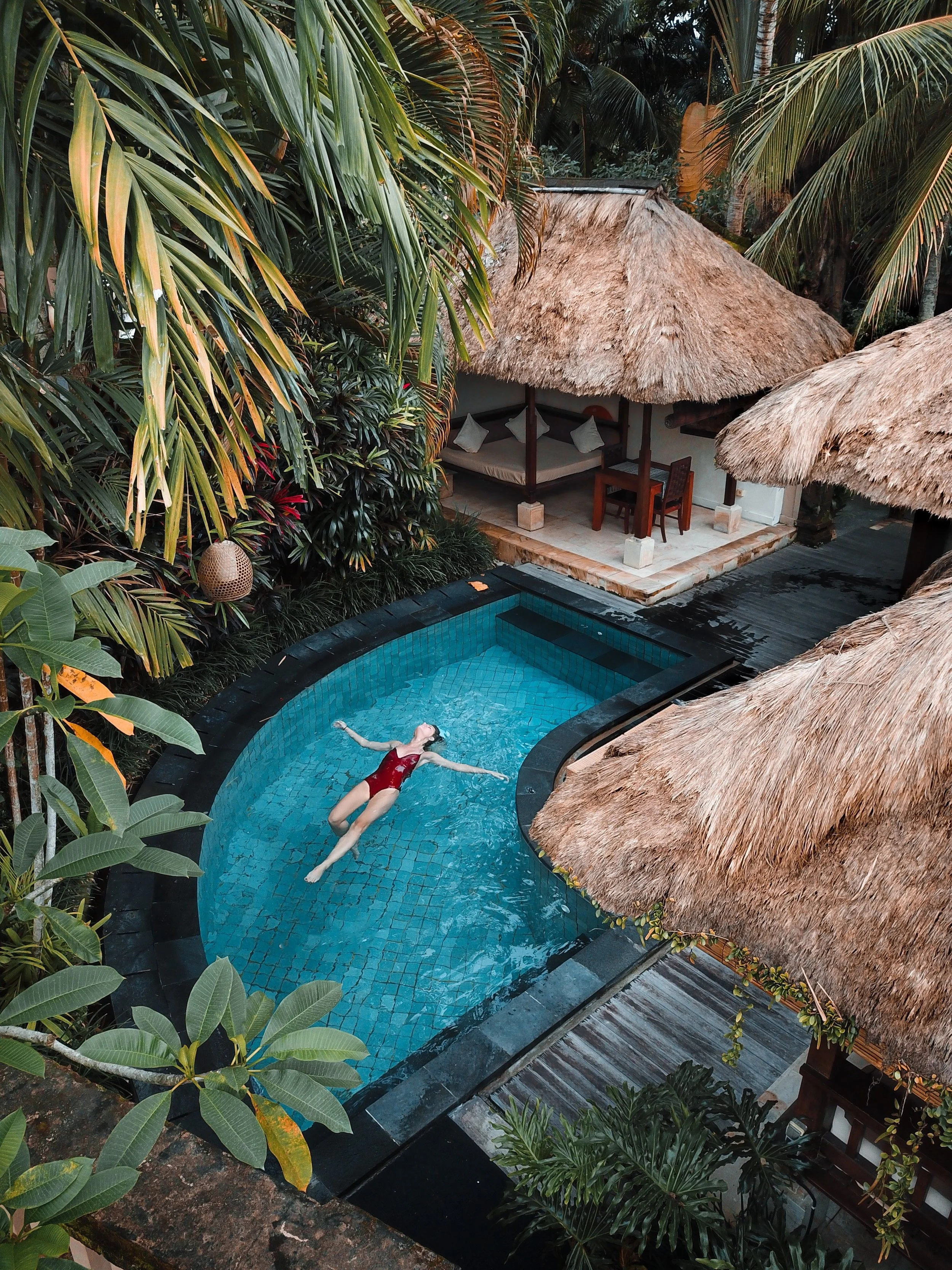 A woman in a red swimsuit is floating in a small, circular swimming pool surrounded by lush tropical plants and thatched-roof structures.