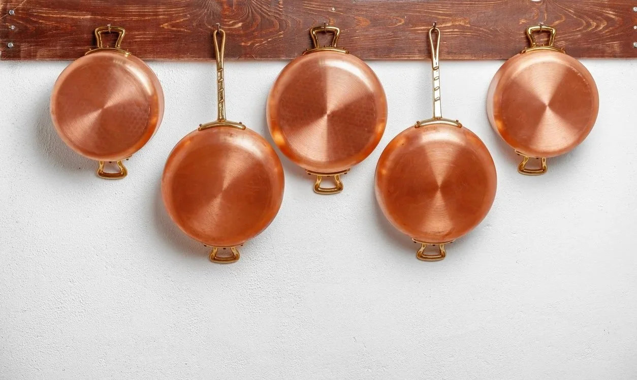 Five copper frying pans hanging in a row from a wooden wall hook.