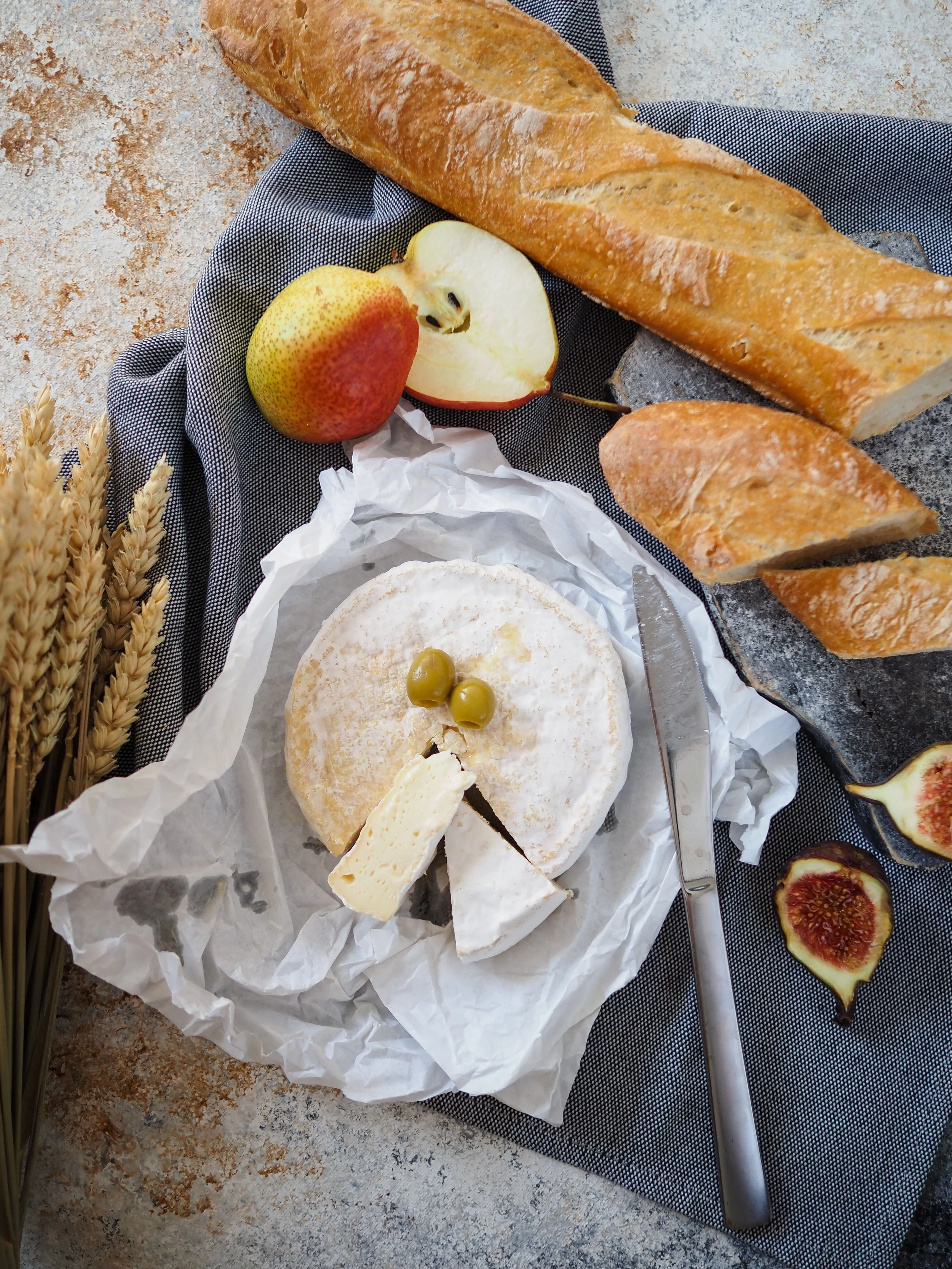 A baguette bread, a sliced apple, a wheel of cheese with a wedge cut out and three green olives on top, a knife, and some slices of figs on a dark cloth on a speckled beige surface.