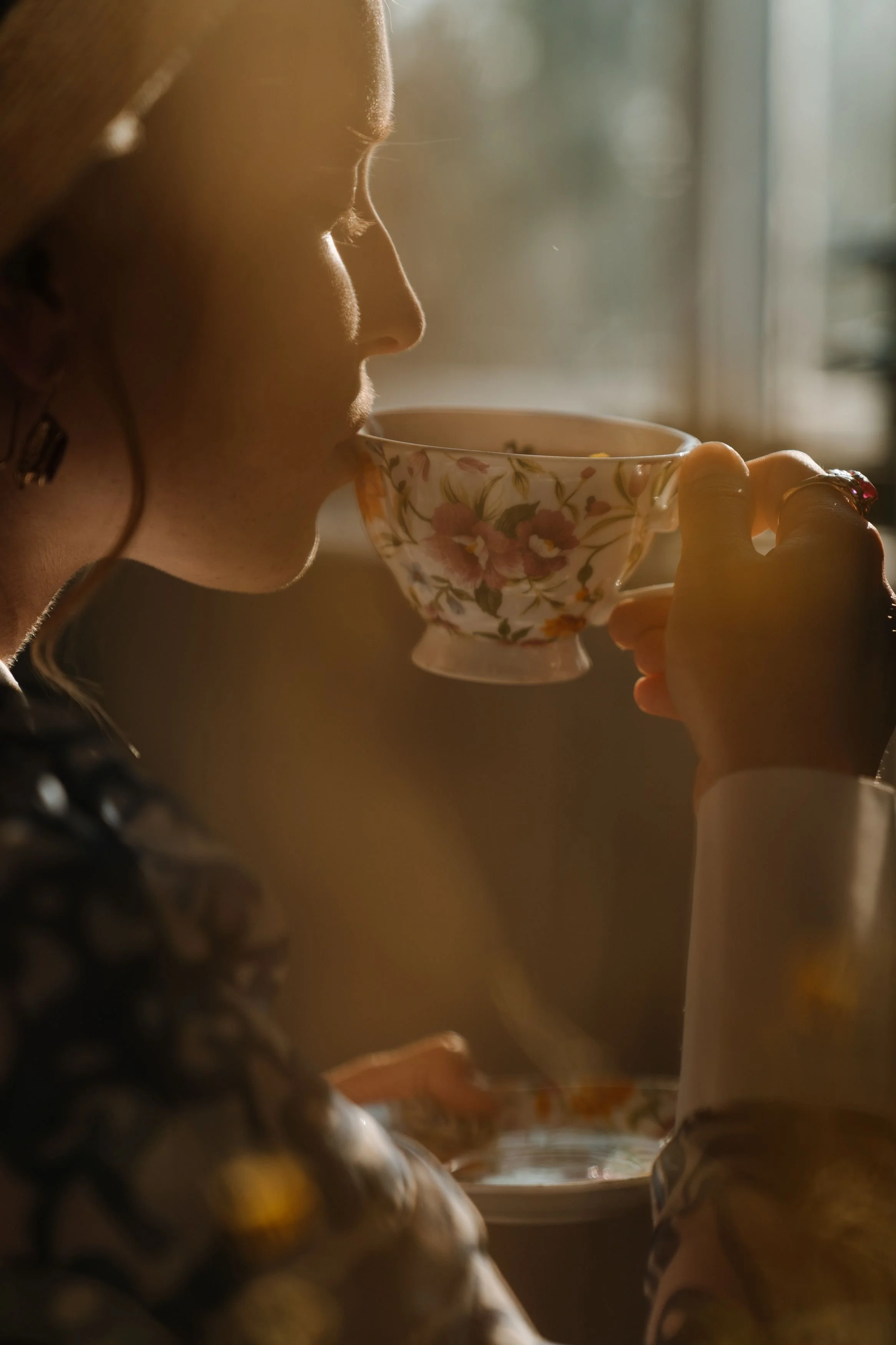 A woman with curly hair drinking from a floral teacup in a softly lit room.