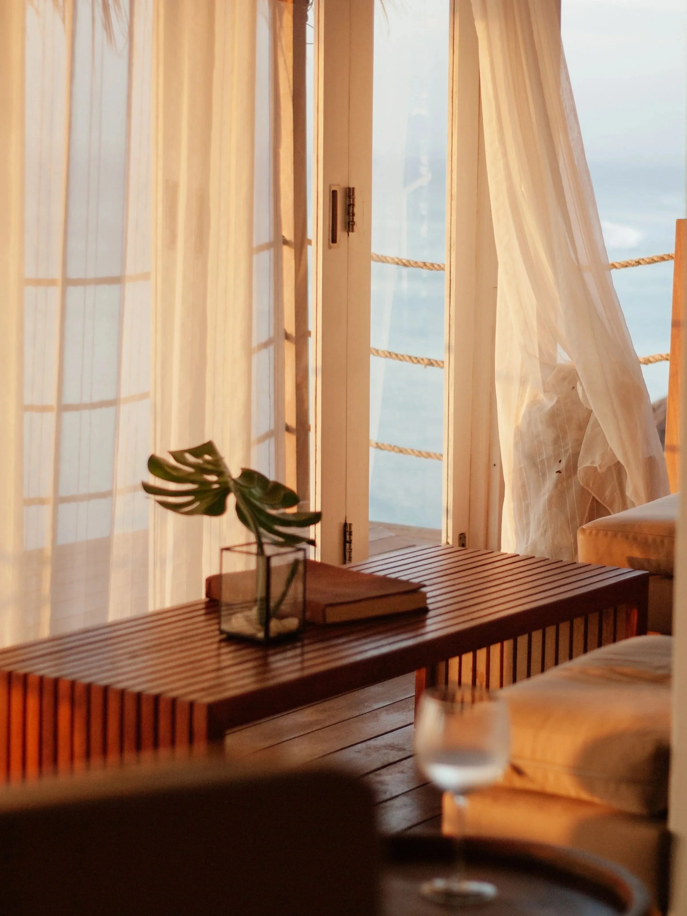 A cozy indoor space with a wooden table holding a glass of wine, a small potted plant, and two books. Behind, there are light-colored sheer curtains and a sliding glass door with a view of the ocean and sky outside.