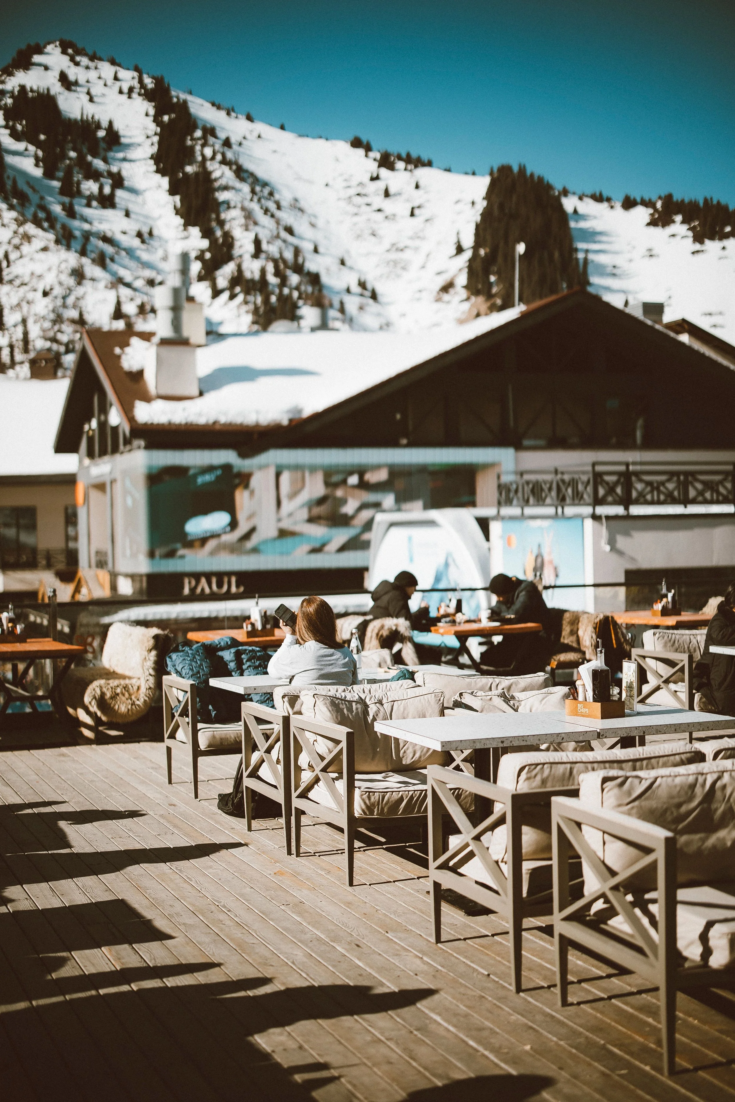 People sitting at outdoor tables on a patio with snow-covered mountains in the background.