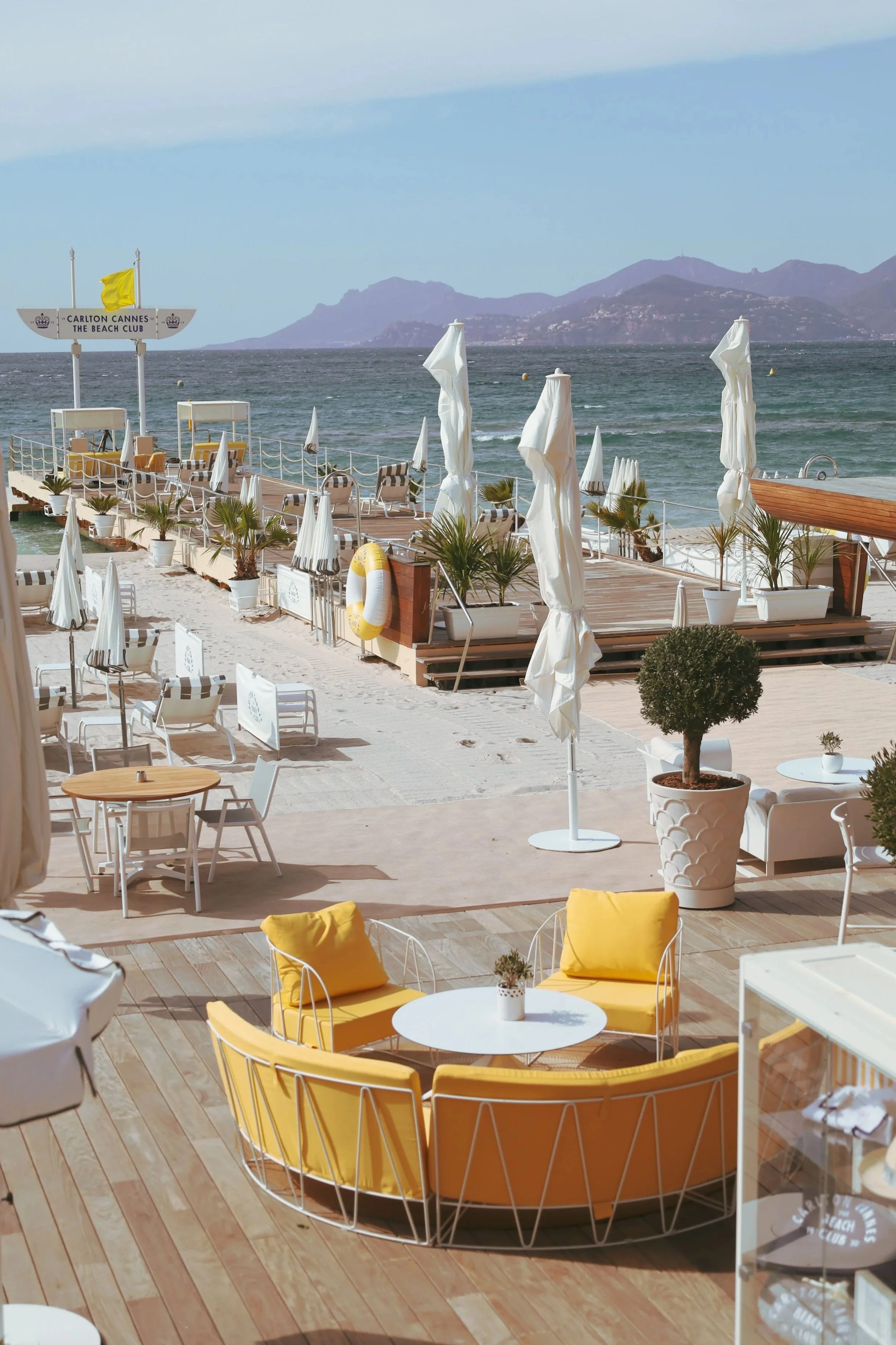 Beachside patio with white and yellow furniture, potted plants, and umbrellas. The ocean and mountains are in the background.