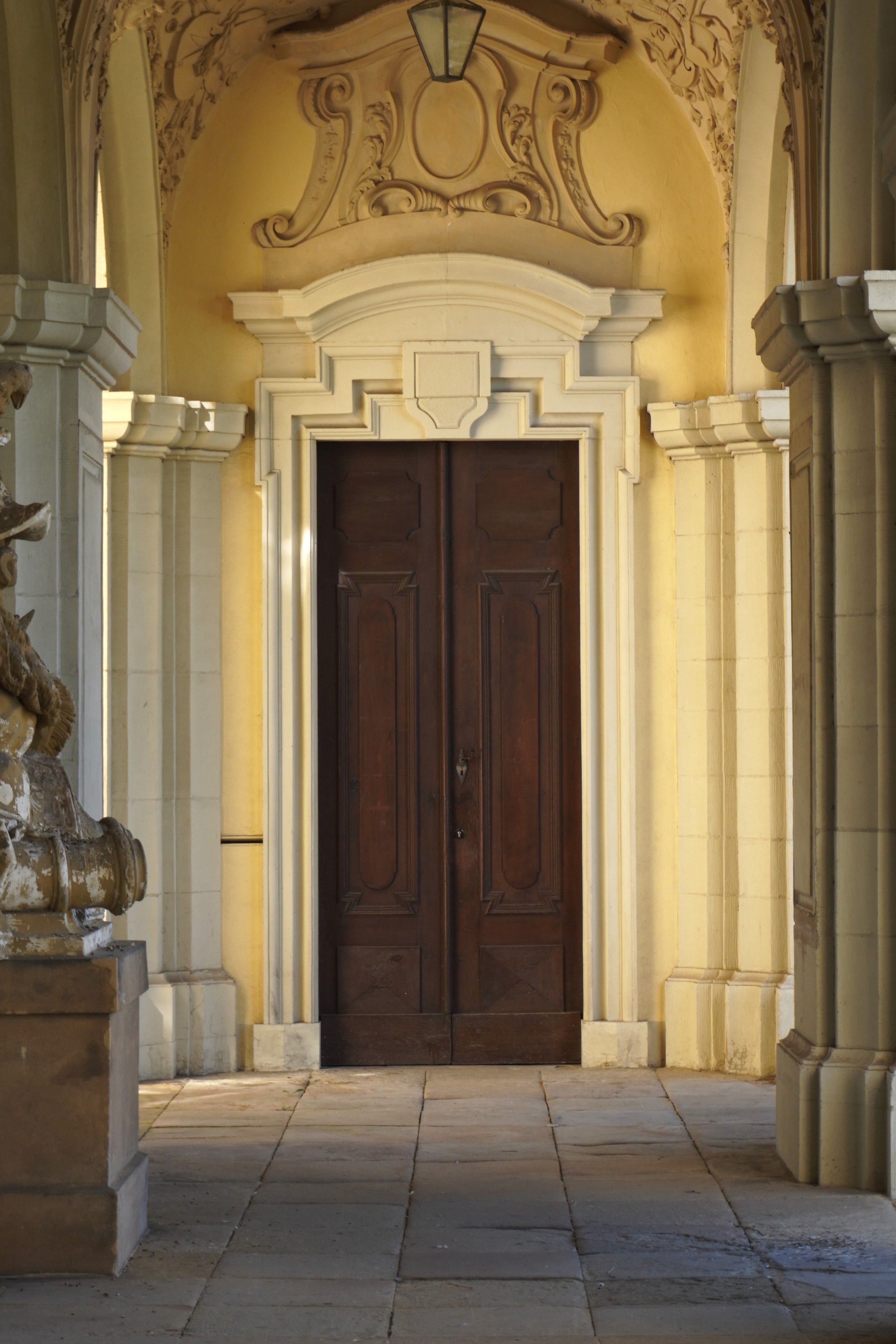 A wooden double door set within a decorative stone archway, flanked by ornate stone columns. The background shows a yellow stucco wall with intricate plasterwork and a hanging lantern ceiling light.