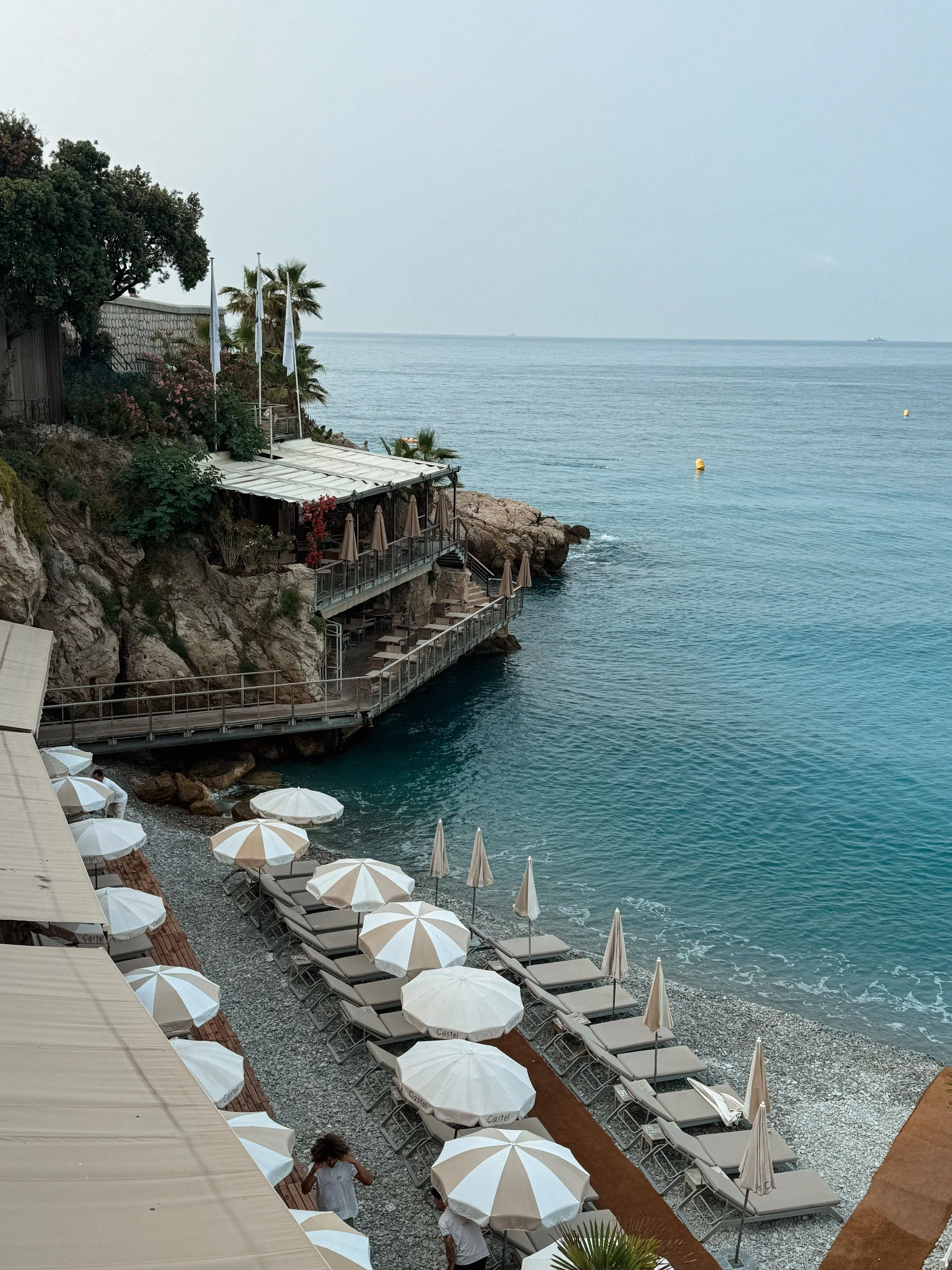 View of a seaside restaurant with white umbrellas and lounge chairs along a rocky beach, overlooking the calm ocean with a ship in the distant horizon.