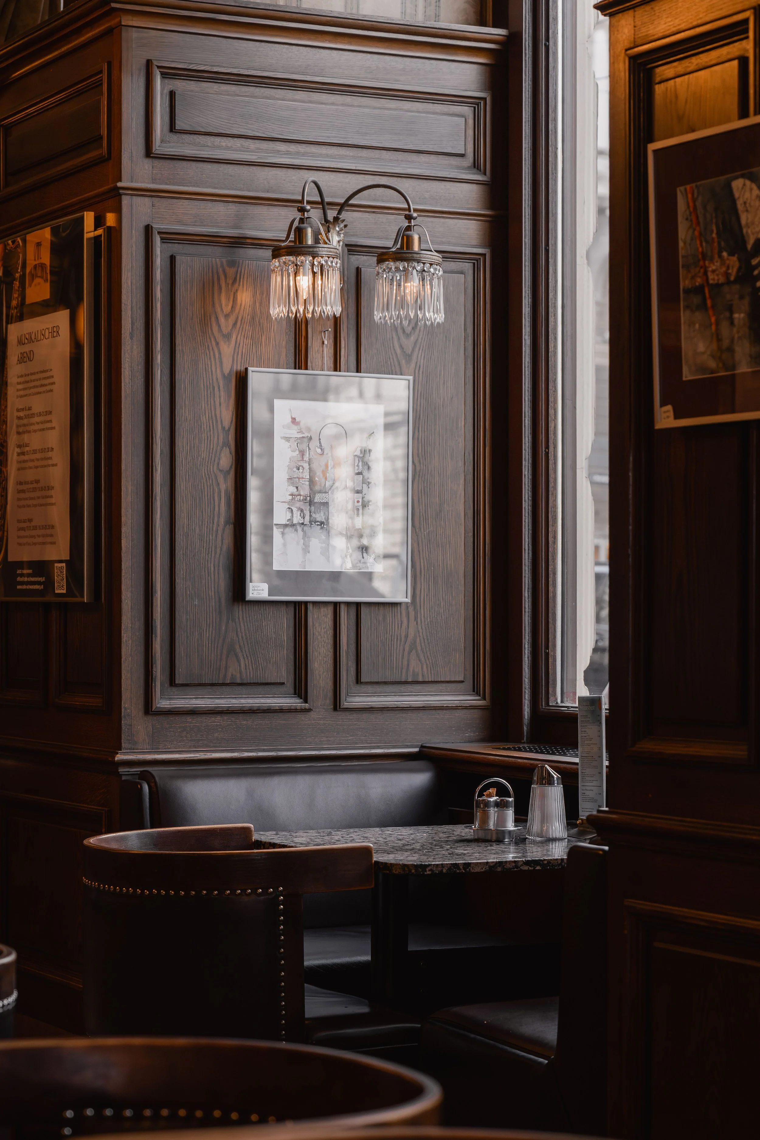 A cozy corner inside a restaurant or cafe with dark wooden paneling, a small round marble table, and chairs. There are framed artwork and posters on the walls, a wall-mounted light fixture with crystal accents, and some table items like glasses and a menu.