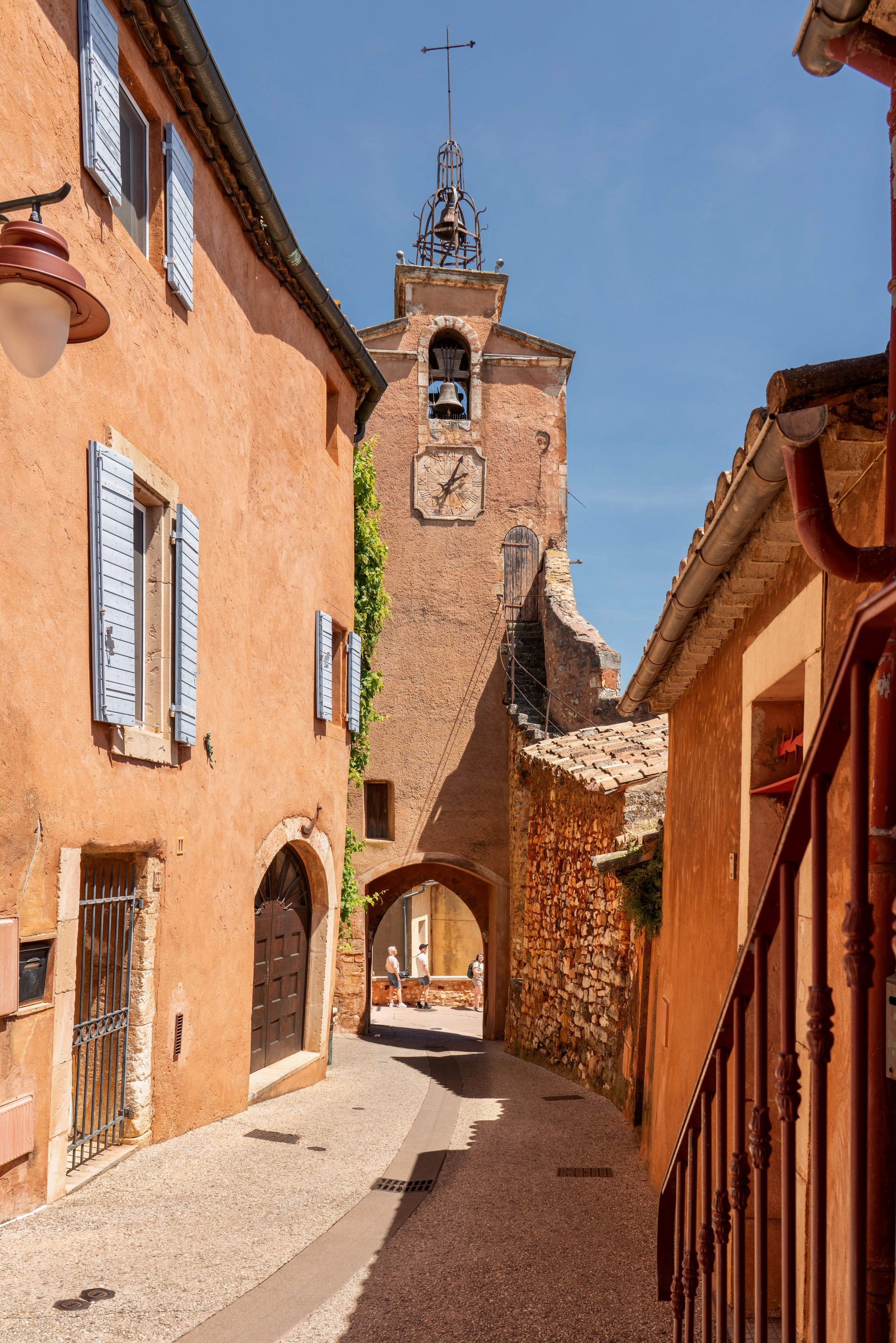 A narrow street in a European town with peach-colored buildings, a church tower with a clock, and a blue sky.