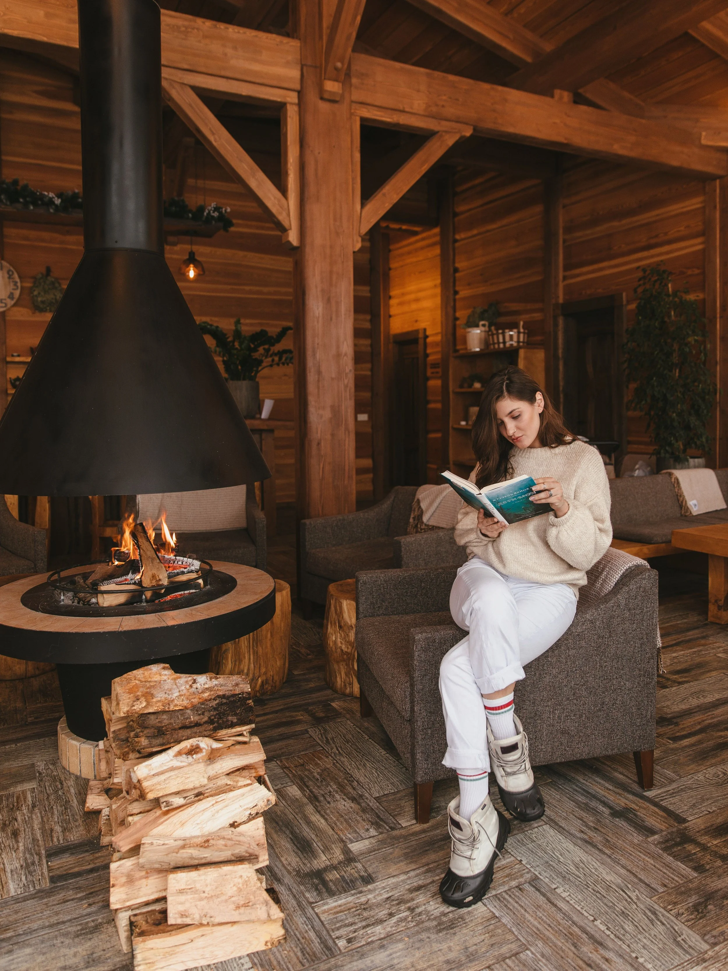 A woman sitting in a cozy cabin interior near a fireplace with wood logs stacked nearby, reading a book. The room features wooden walls, beams, and warm lighting, creating a rustic atmosphere.