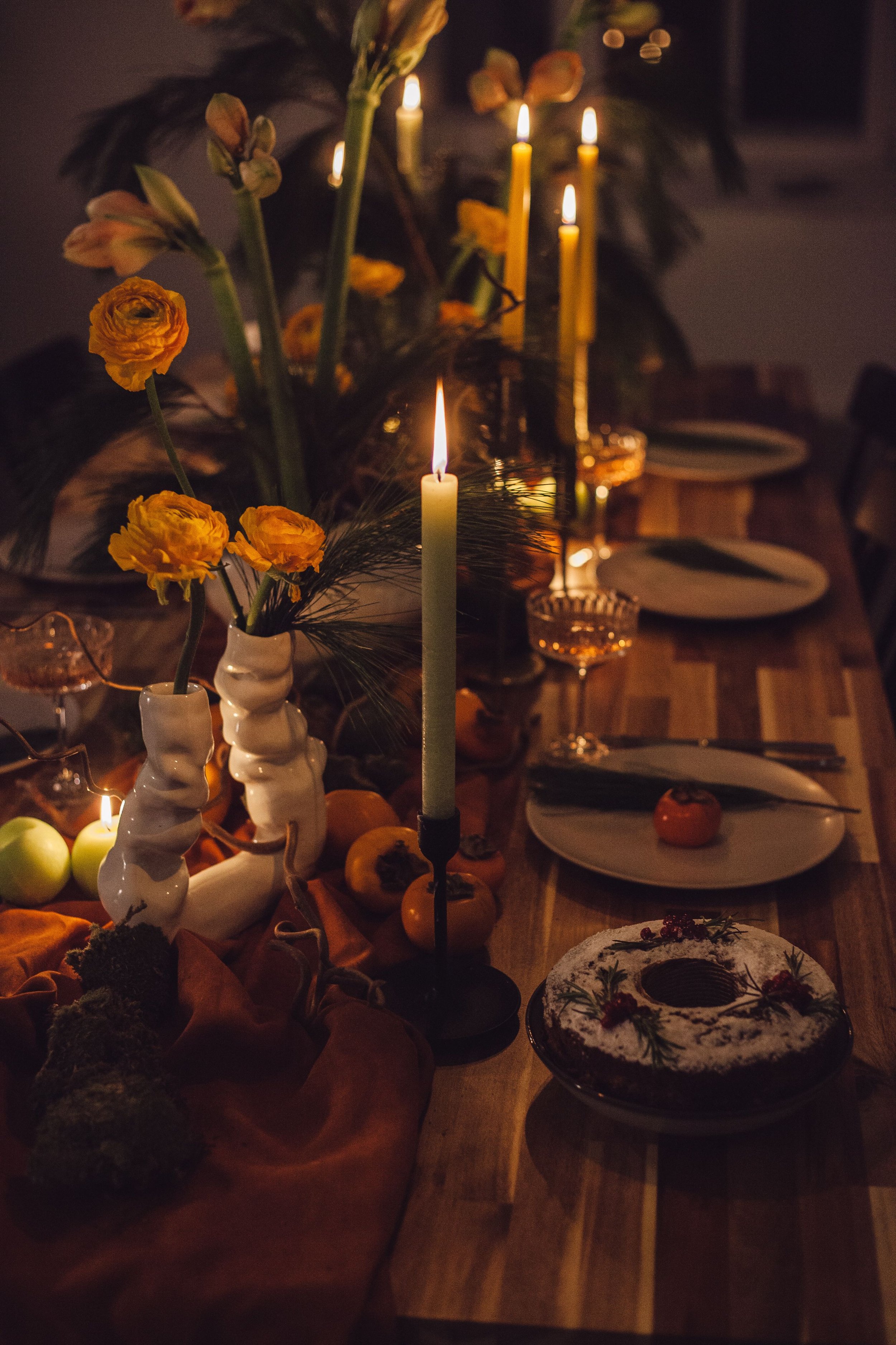 A dimly lit dinner table with candles, flowers, and assorted food items including a chocolate cake with berries and herbs.