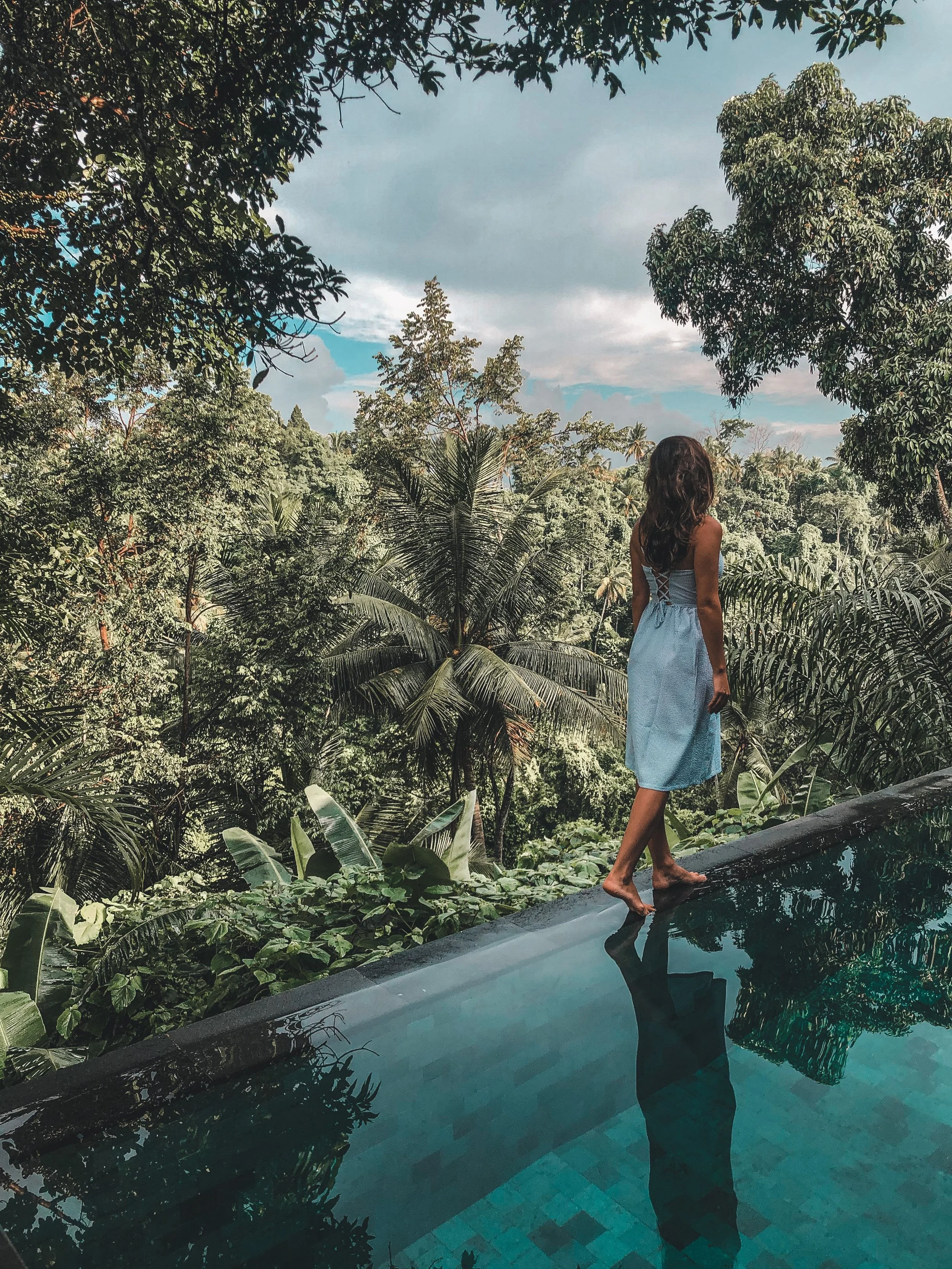 A woman stands on the edge of a swimming pool, overlooking a lush, green jungle landscape. She is barefoot, wearing a light blue dress, and her reflection is visible in the water. The scene is serene with a cloudy sky overhead.