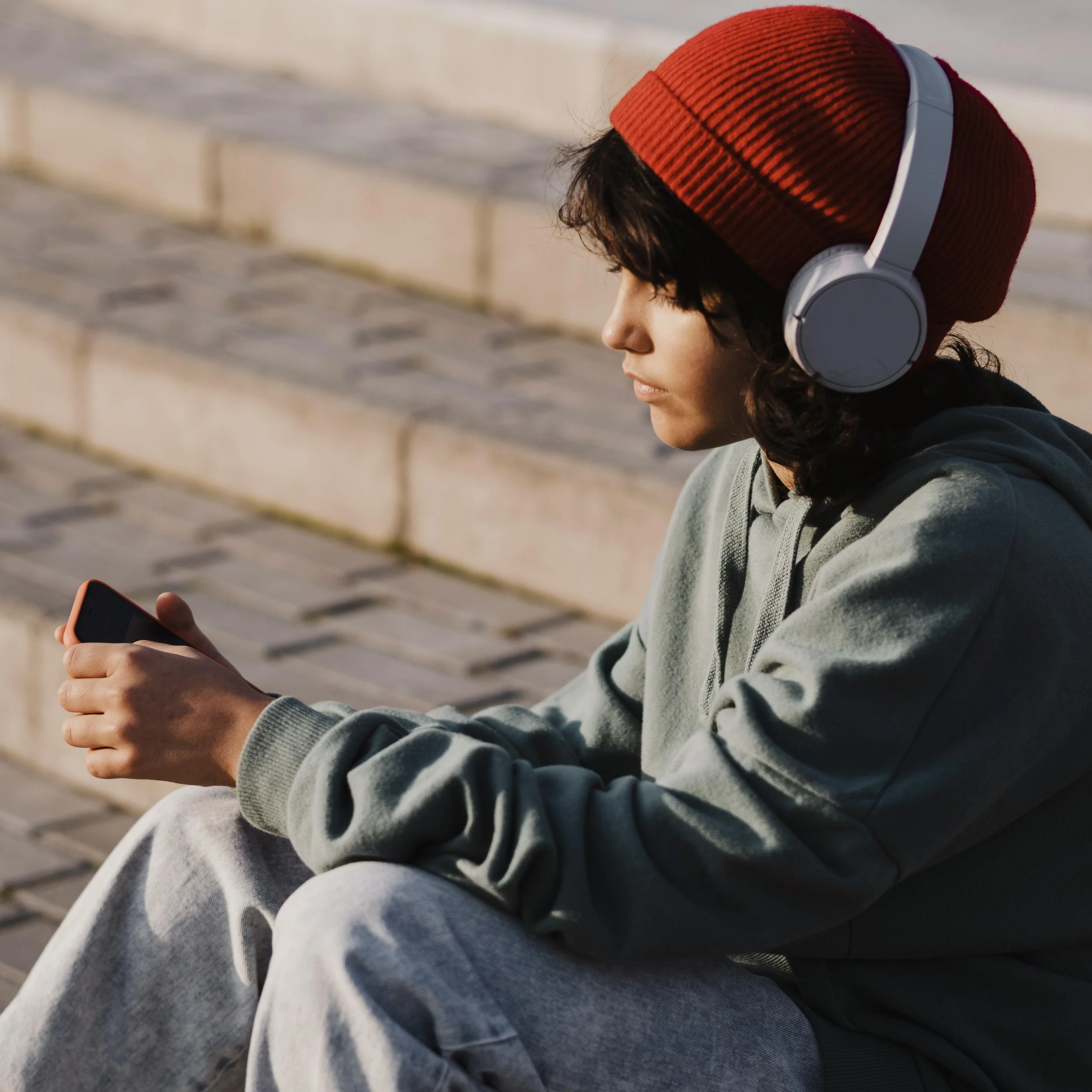 A person sitting on outdoor steps, wearing a red beanie and headphones, looking at their phone.