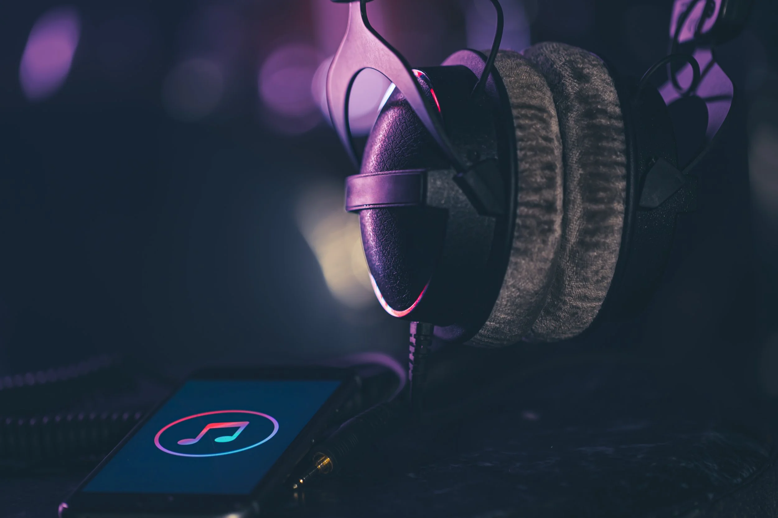 A pair of over-ear headphones hanging on a hook, with a smartphone displaying a music note icon nearby, in a low-light setting with purple and blue hues.