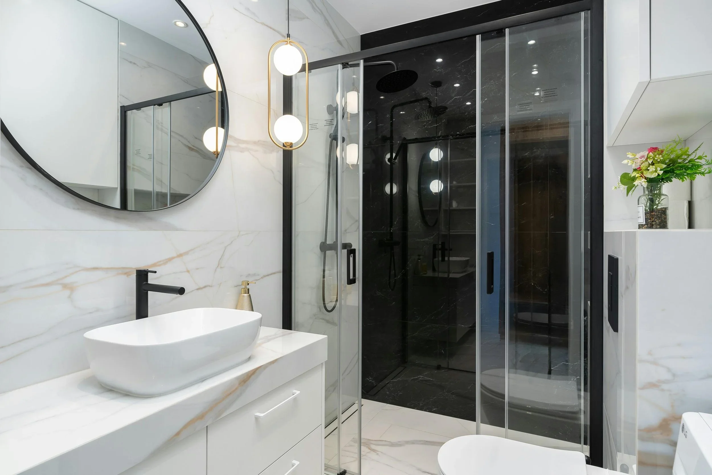 Modern bathroom with white marble walls and a black shower enclosure. Includes a white vessel sink with a black faucet and a round mirror, contemporary lighting fixtures, and a small vase with flowers on a shelf.