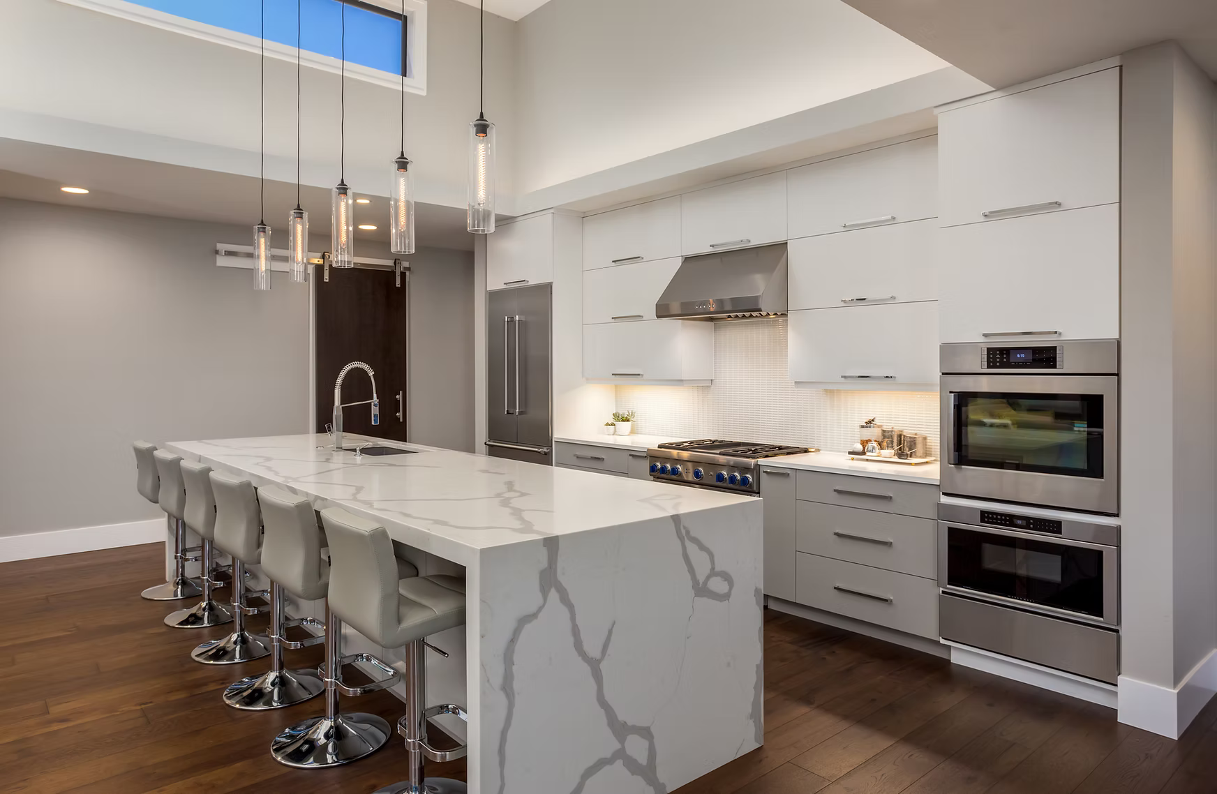 Modern kitchen with white cabinets, stainless steel appliances, a large marble island with bar stools, pendant lights, and hardwood floors.