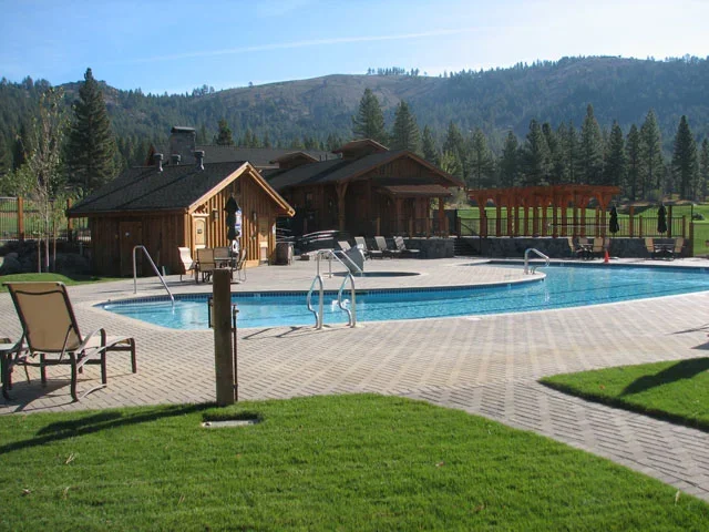 A backyard swimming pool with lounge chairs, surrounded by grassy area and a paved patio, with wooden buildings and lush green mountains in the background.