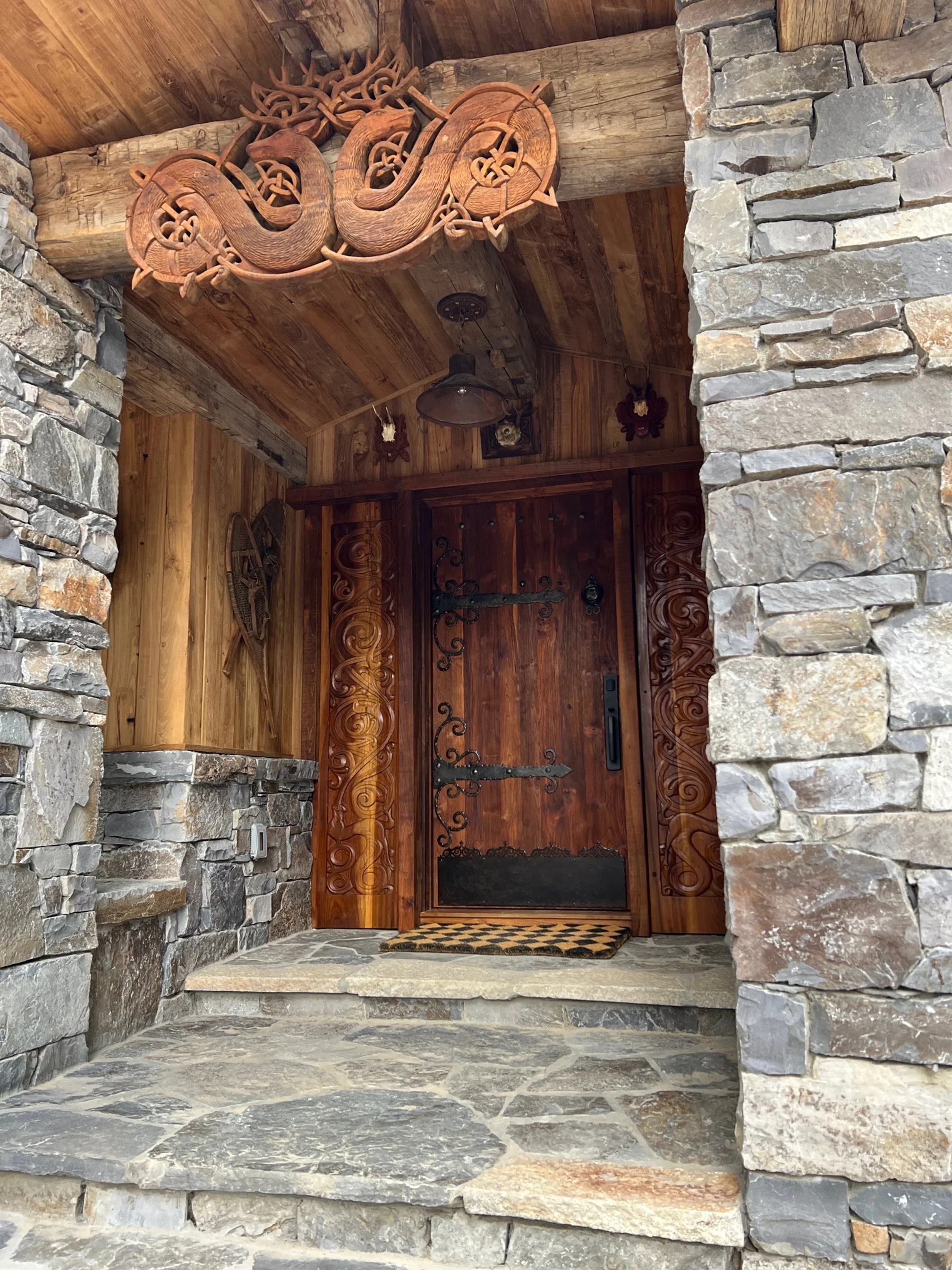 Wooden front door with black wrought iron hardware, flanked by stone walls and a stone step, with carved wood accents and decorative metal and wooden elements in a rustic lodge-style entrance.