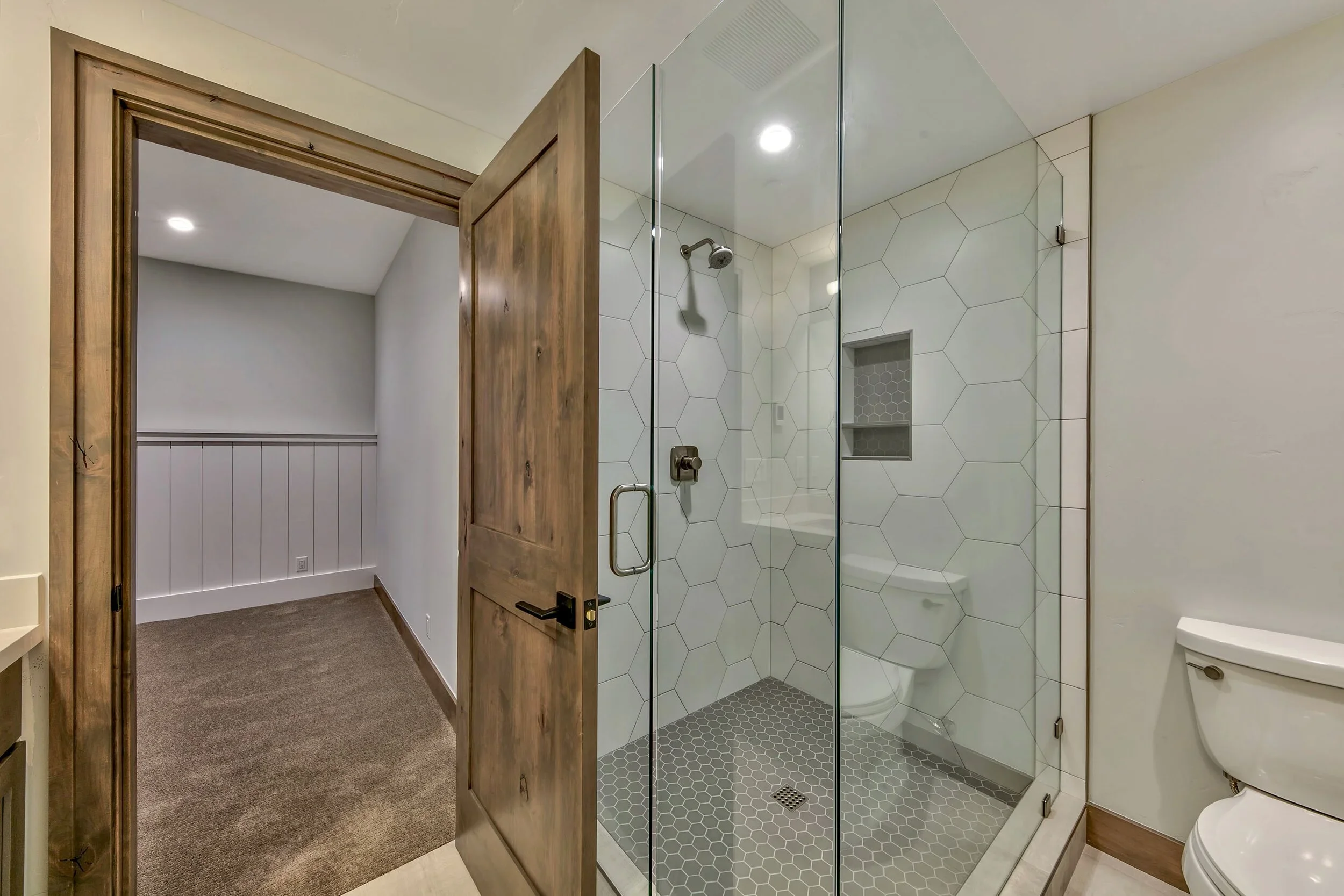 Bathroom with a glass-enclosed shower, white hexagonal tiles, a built-in niche, a wall-mounted shower head, a toilet, and a partially open wooden door leading to a room with white wainscoting and brown carpet.