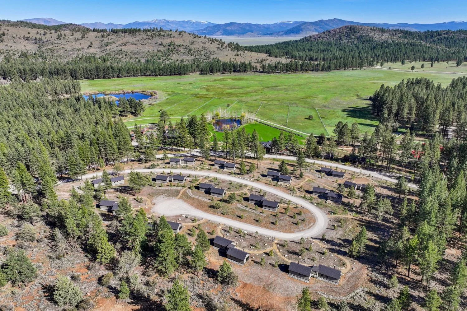 Aerial view of private cabins arranged along winding hillside roads overlooking pine forests, expansive green meadows, two ponds, and distant mountains.