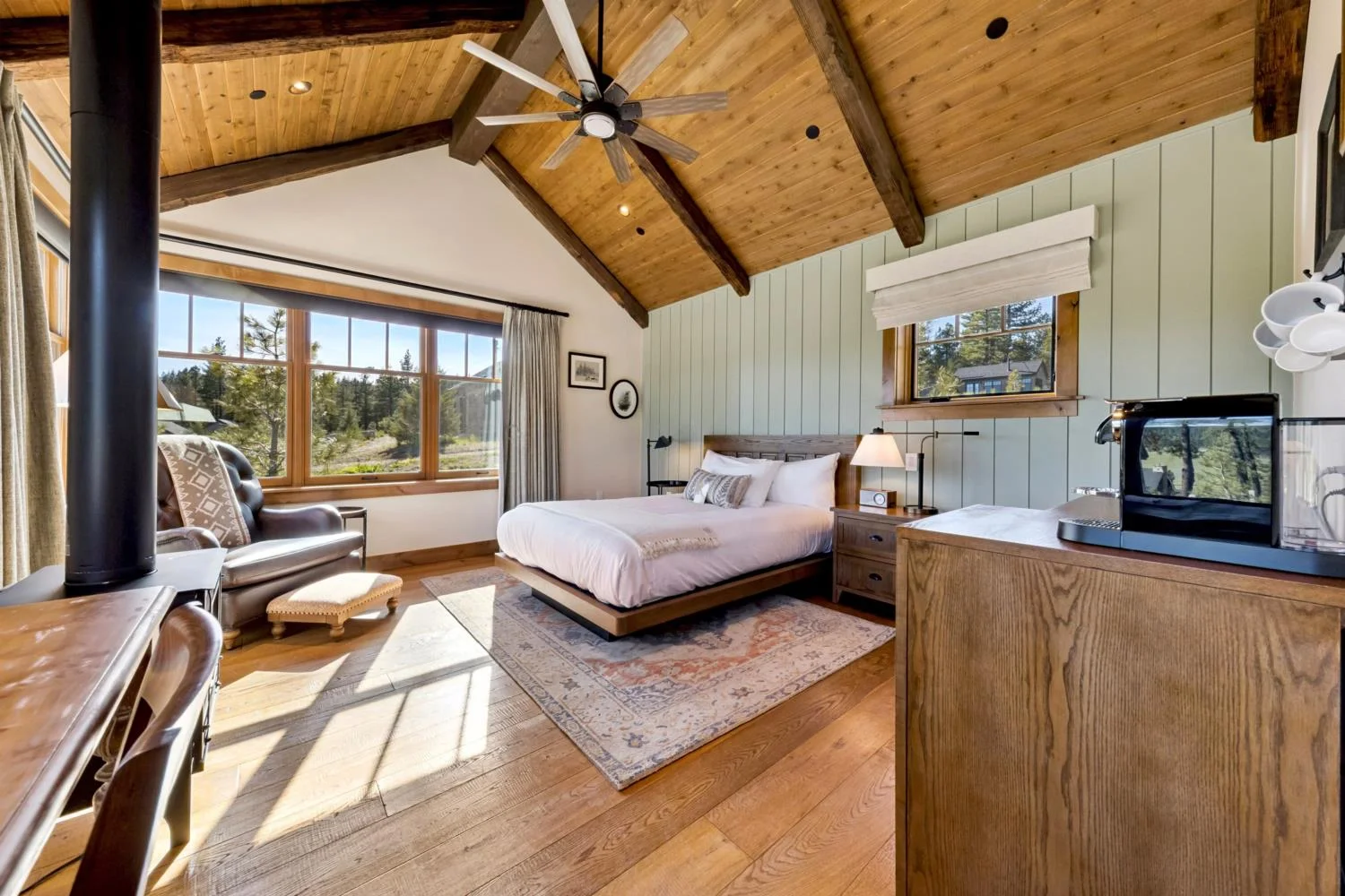 Private cabin bedroom with a vaulted wood ceiling, exposed beams, sage green paneled walls, natural wood flooring, a freestanding stove, coffee station, and large windows overlooking the forest.