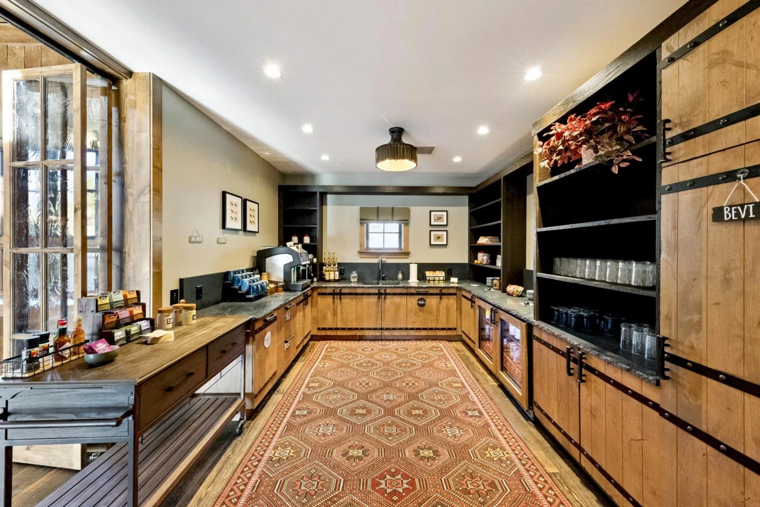 Lodge beverage station with natural wood cabinetry, black countertops, open shelving, coffee service, undercounter refrigerators, a small sink, and a long patterned rug.