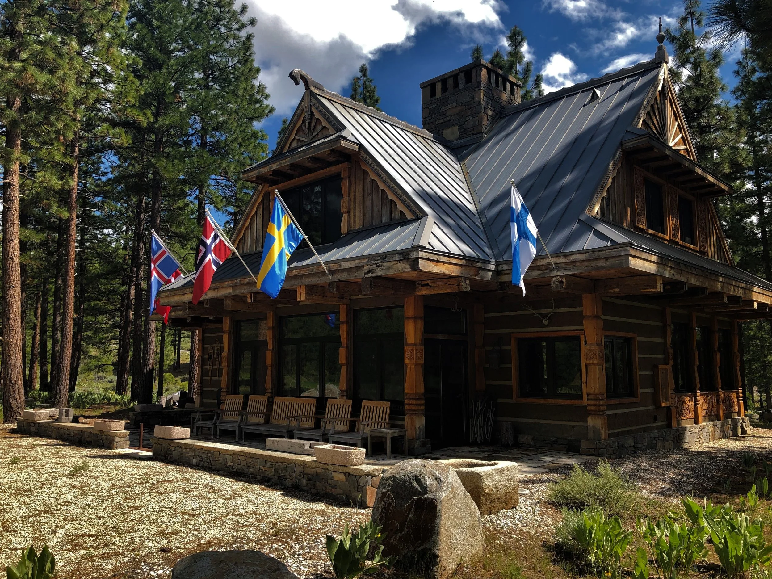 A rustic wooden house with a metal roof, surrounded by tall pine trees, displays several flags including Scandinavian flags on a porch with benches.