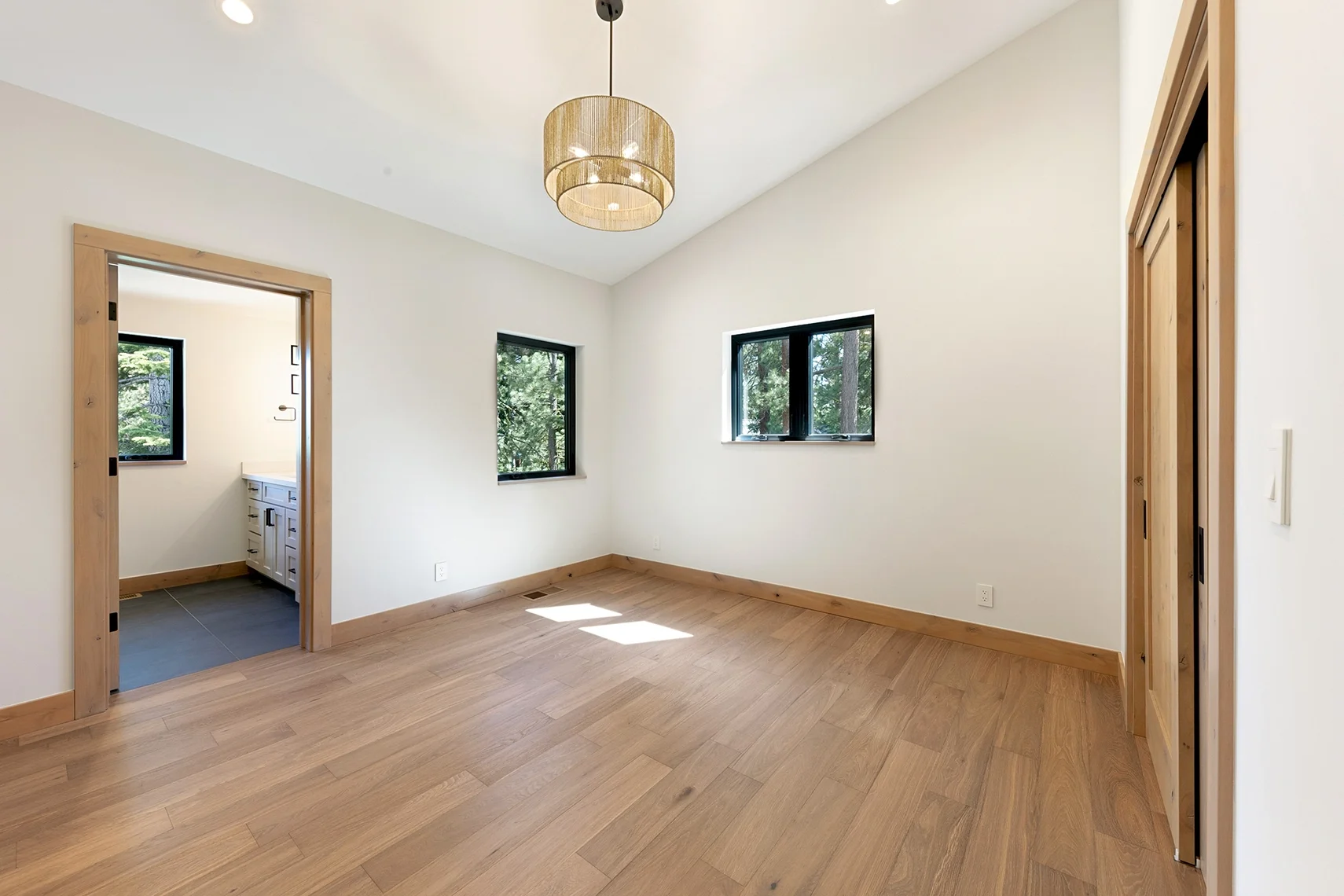 Bright modern bedroom with white walls, natural wood flooring, two windows, a pendant light, and an attached bathroom with black tile flooring.