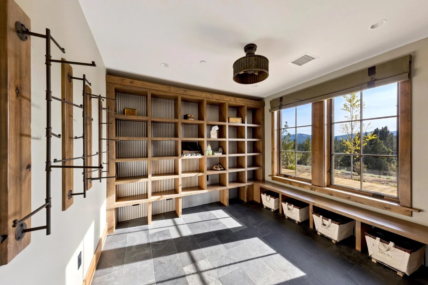 Lodge gear room with built-in wood cubbies, wall-mounted drying racks, a storage bench with baskets, dark slate tile flooring, and large windows overlooking the mountain landscape.