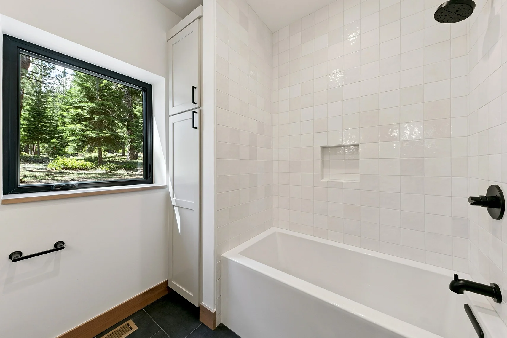 Bright white bathroom with a tiled bathtub, clean modern finishes, and a window overlooking a peaceful pine tree landscape.