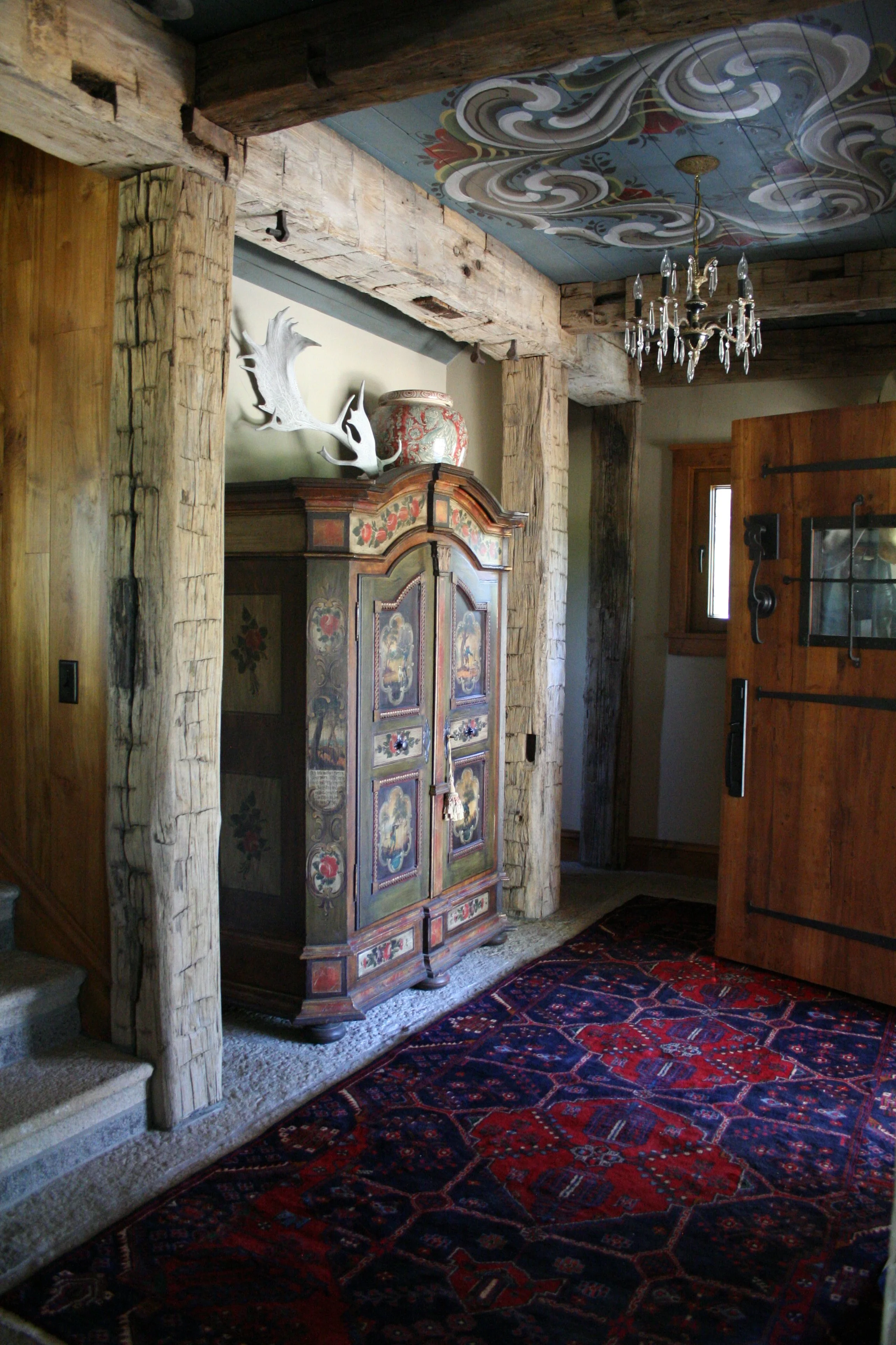 Interior of a rustic room with wooden beams, a chandelier, an ornate armoire, a small window, and a decorative ceiling painted with clouds and swirls. A red patterned rug covers the floor.