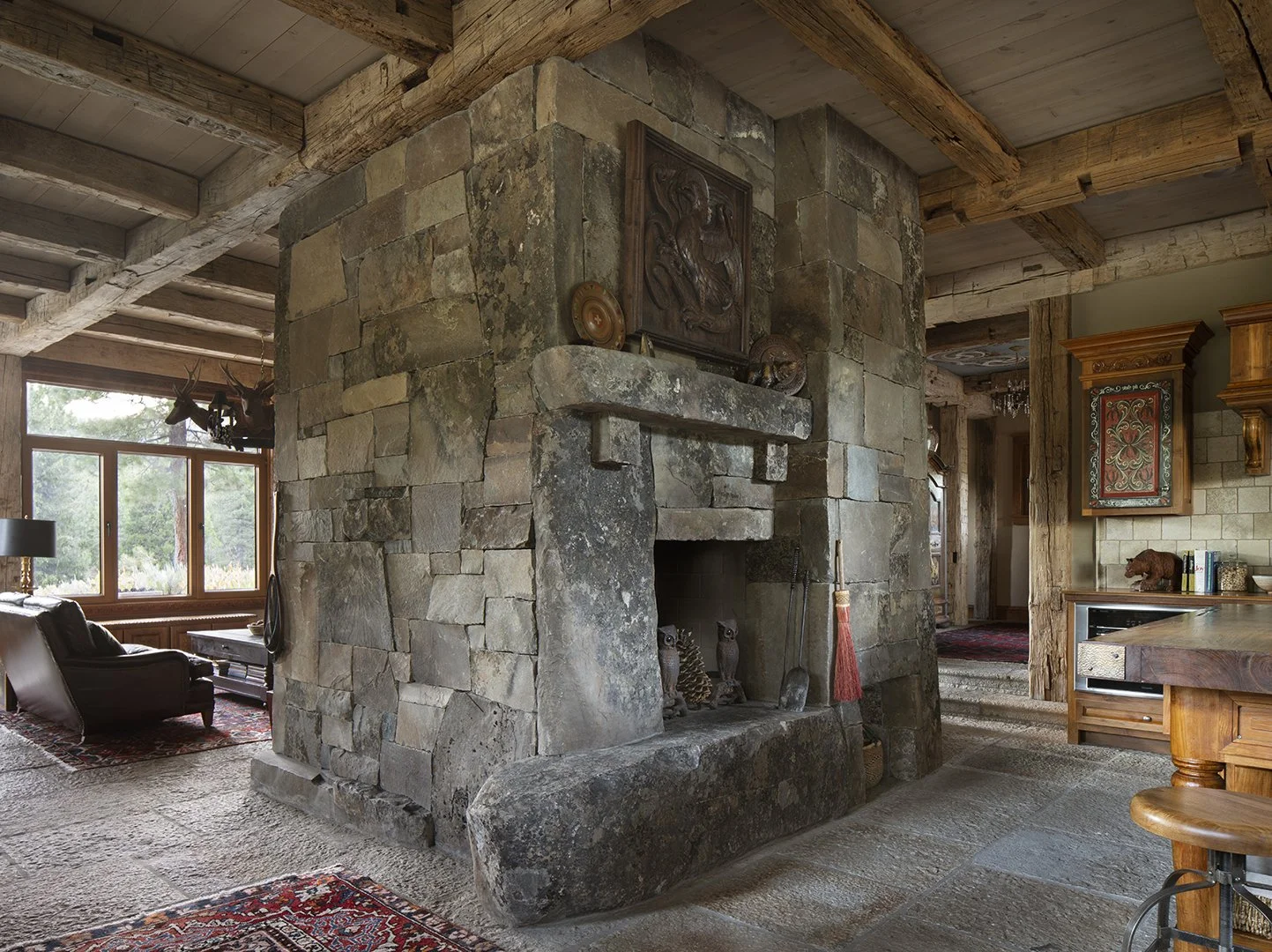 Interior of a rustic living room featuring a large stone fireplace with wooden beams on the ceiling, a window with outdoor greenery view, a leather armchair, and decorative items on the mantel.