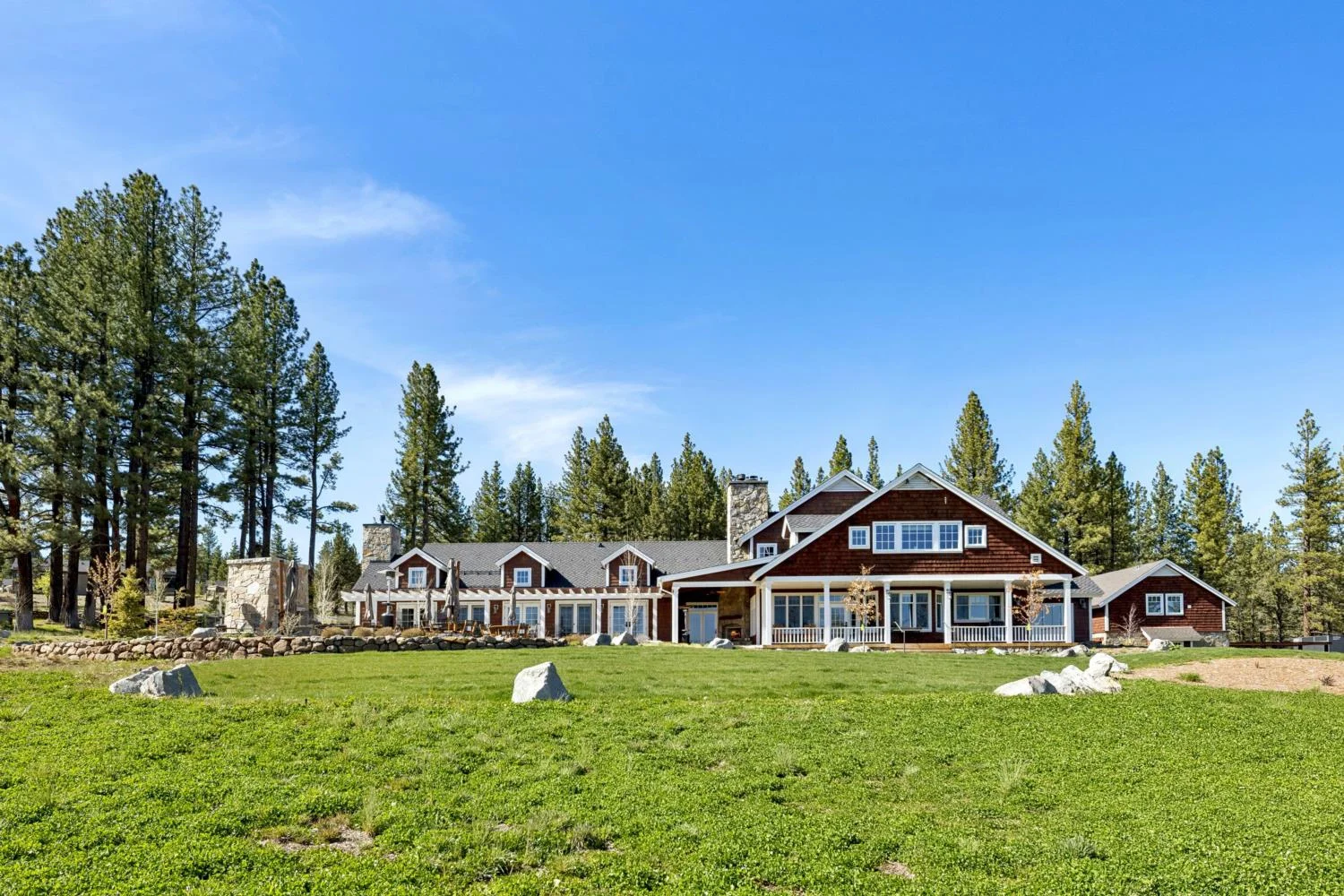Large ranch-style lodge with dark wood siding, white trim, covered porches, and stone chimneys overlooking a green meadow beneath a clear blue sky.