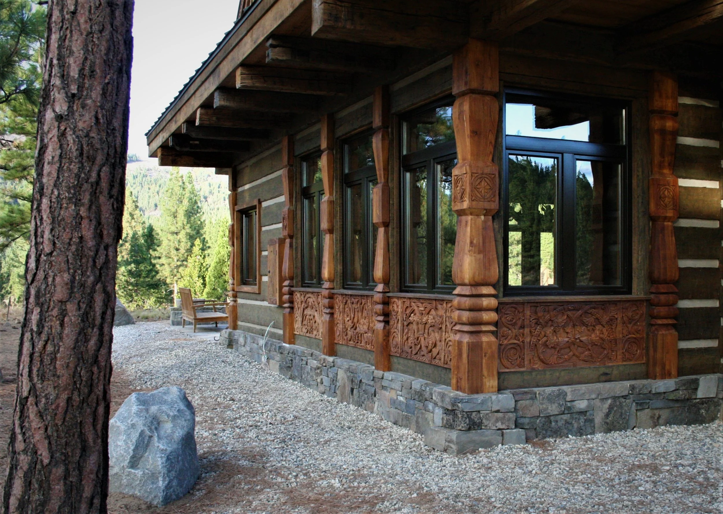 Image of a rustic log cabin with intricate wooden carvings and large glass windows, surrounded by pine trees and a gravel walkway.