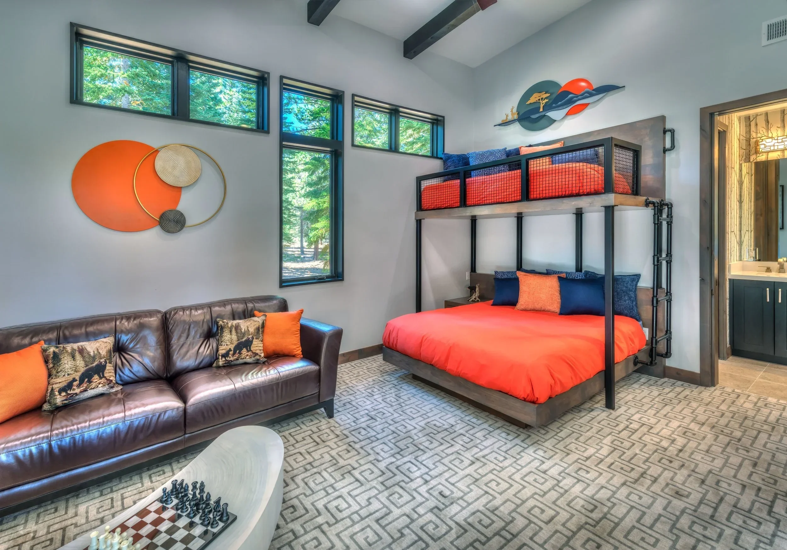 Room with a brown leather sofa with orange and bear-themed pillows, a bunk bed with orange bedding, and abstract wall art, with large windows showing green trees outside.