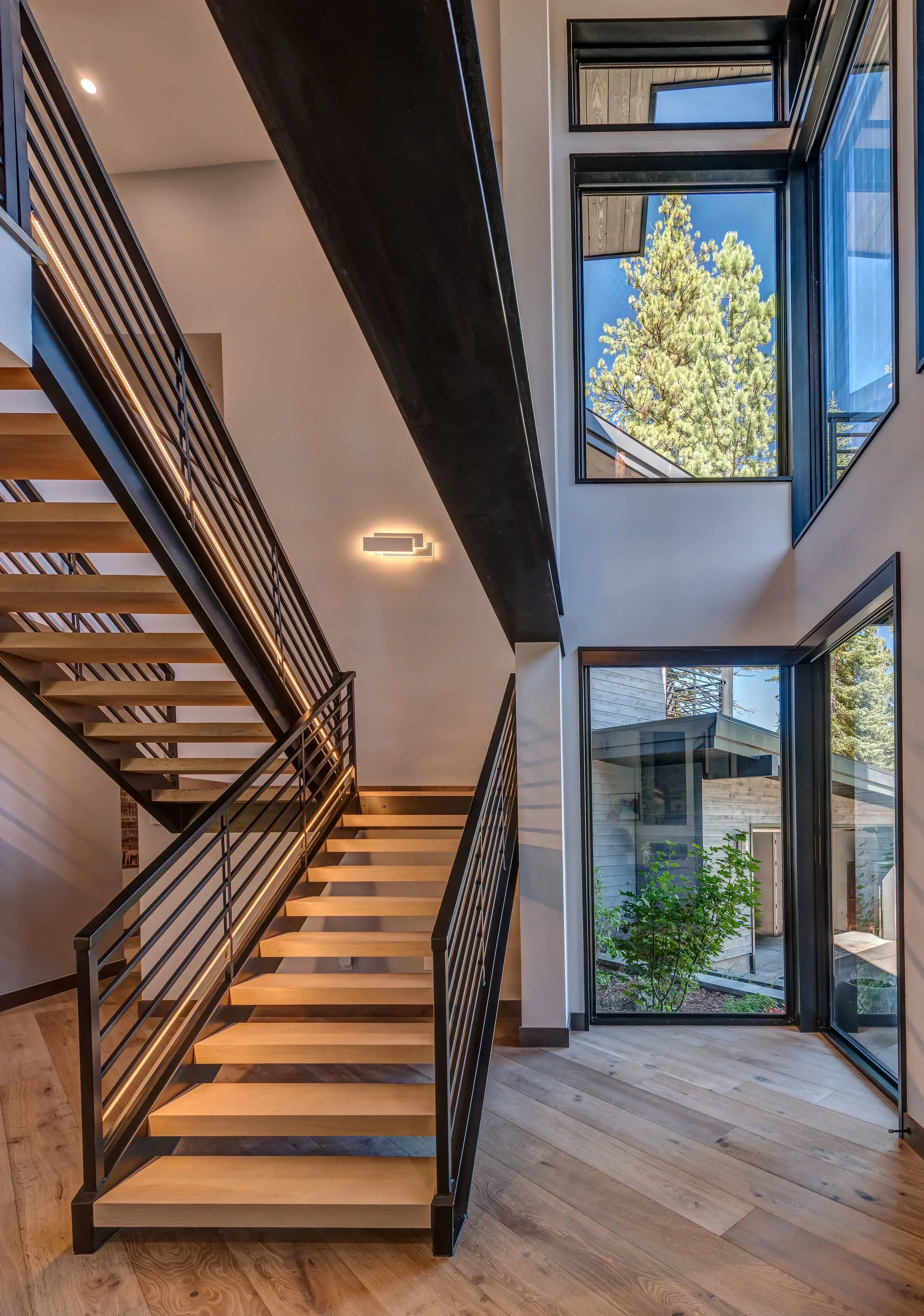 Modern interior staircase with wooden steps and black metal railings in a house with large windows showing trees outside.
