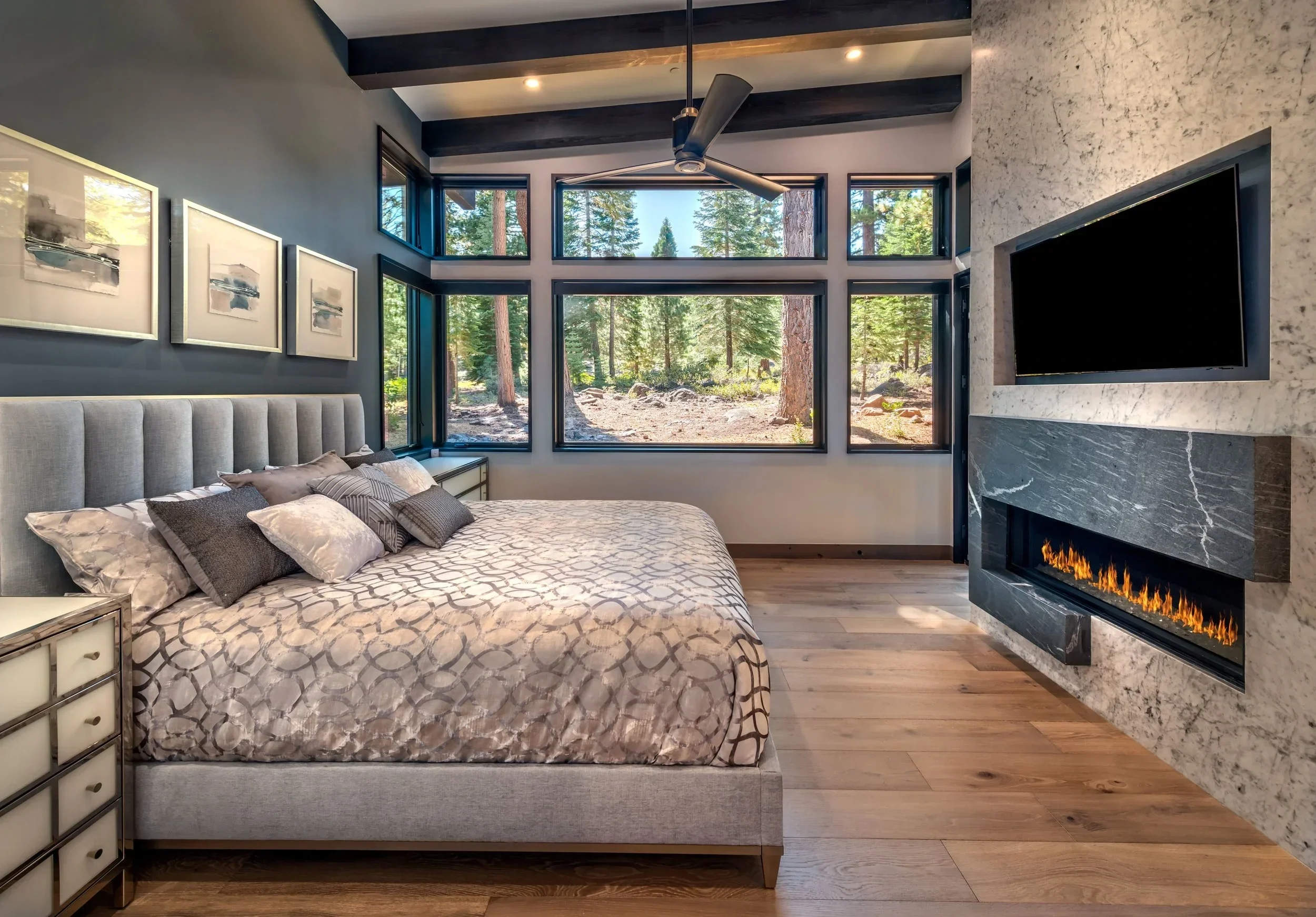 Modern bedroom with large windows showing a forest view, a bed with a patterned duvet and multiple pillows, a wall-mounted flat-screen TV, and a fireplace.
