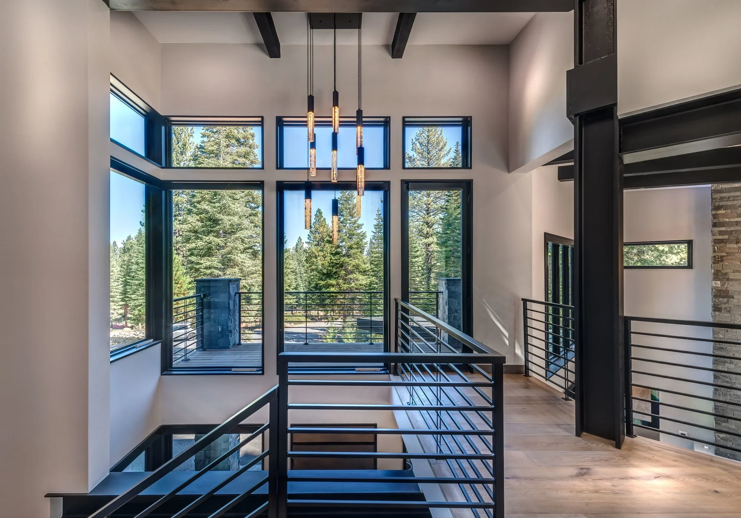 Interior view of a modern house with large floor-to-ceiling windows overlooking a forest, featuring black metal railings, a high ceiling, and a minimalist chandelier with multiple hanging lights.